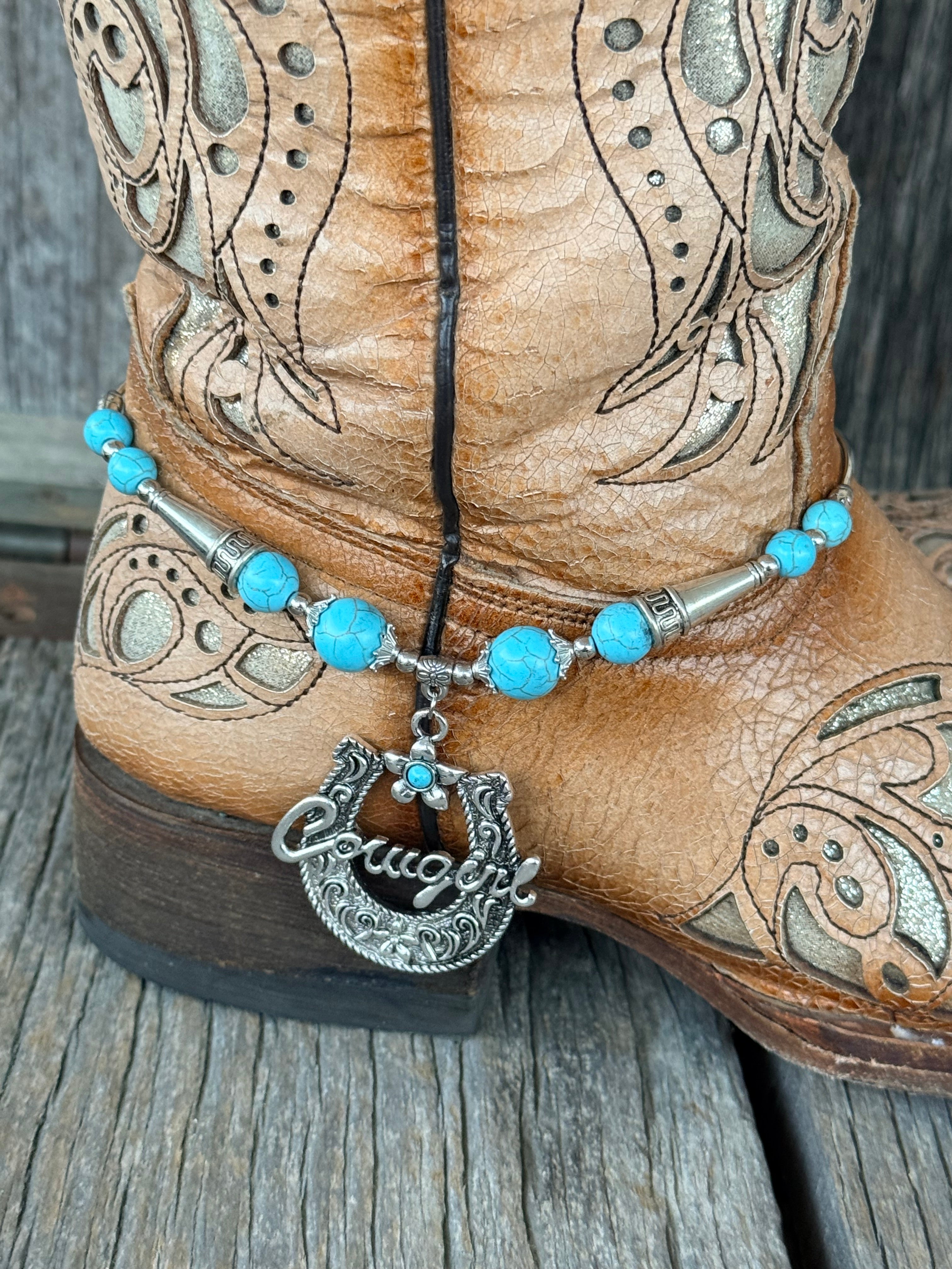 Close-up of turquoise round bead and silver tube spacer boot bracelet with large ornate silver horseshoe Cowgirl script pendant and turquoise flower charm drop at heel of tan tooled cowboy boot
