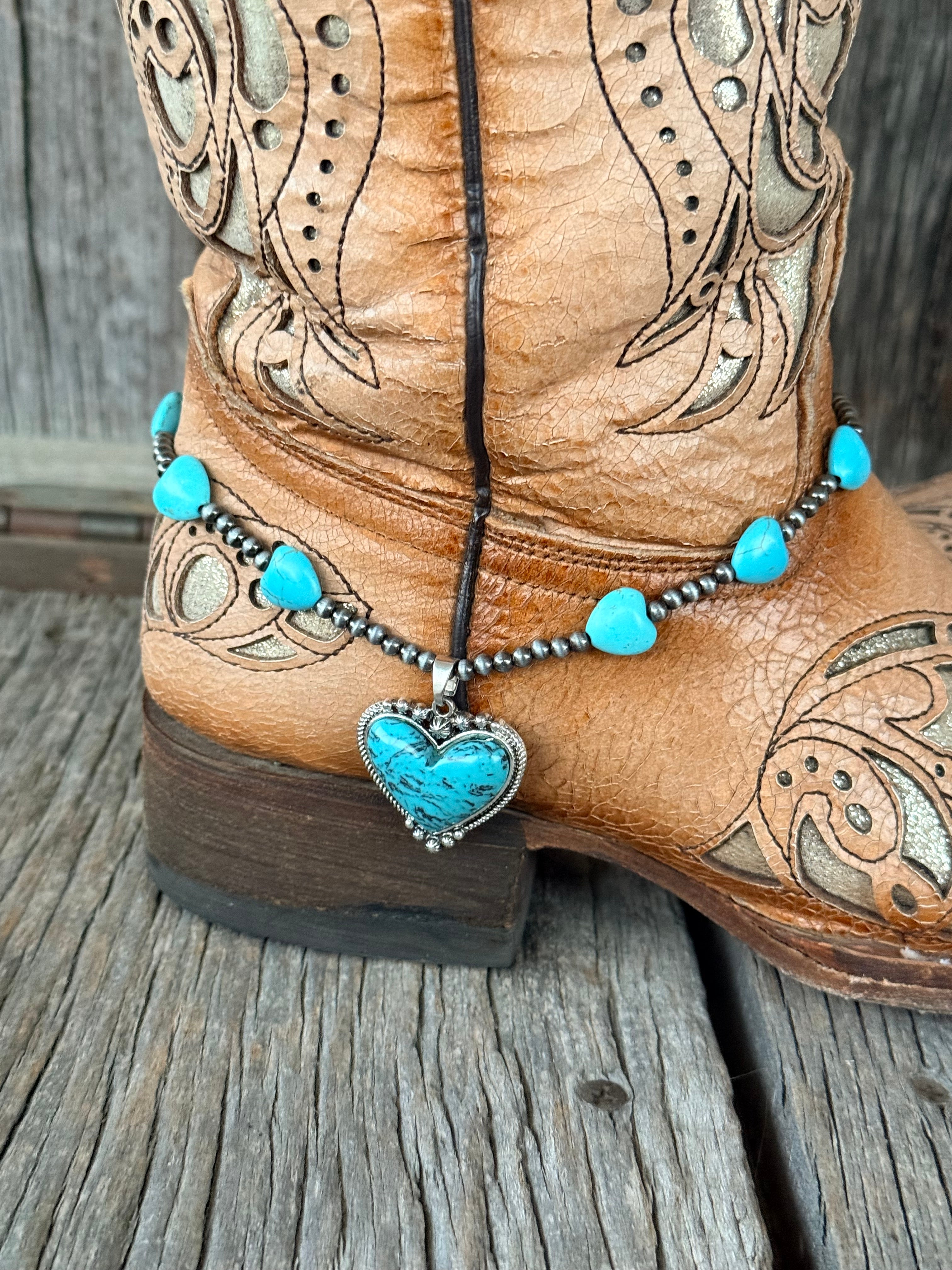 Close-up of turquoise heart bead and Navajo pearl gunmetal bead boot bracelet with large turquoise heart stone pendant in rope-twist silver setting at heel of tan tooled cowboy boot
