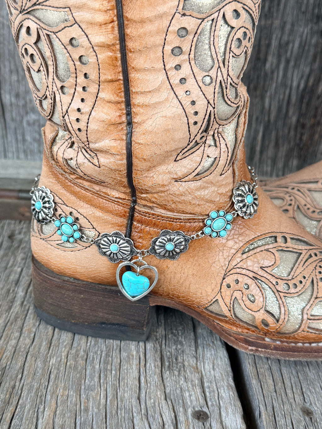 Close-up of linked silver scalloped flower concho chain boot bracelet with turquoise cabochon centers and large open silver heart frame with turquoise stone drop at heel of tan tooled cowboy boot
