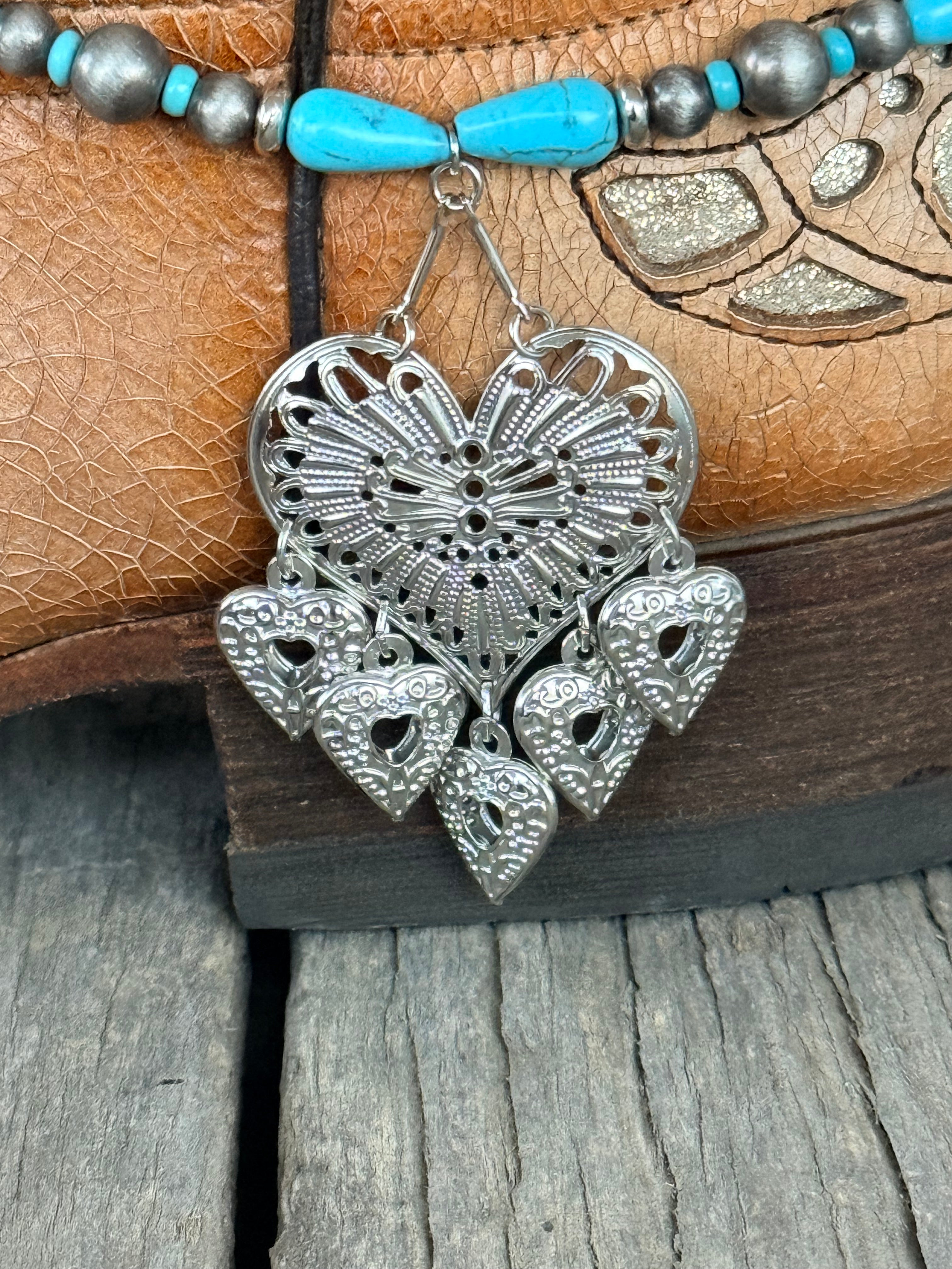 Detail close-up of large ornate silver filigree heart pendant with open lace-cut sunburst design and five dangling silver heart charms on turquoise teardrop and Navajo pearl boot bracelet
