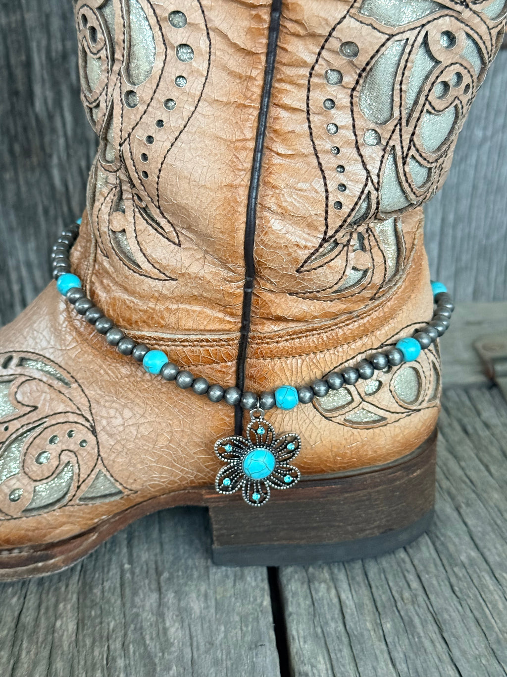 Side view of gunmetal Navajo pearl and turquoise bead boot bracelet with ornate silver flower concho pendant draping at ankle of tan tooled laser-cut cowboy boot on wood surface

