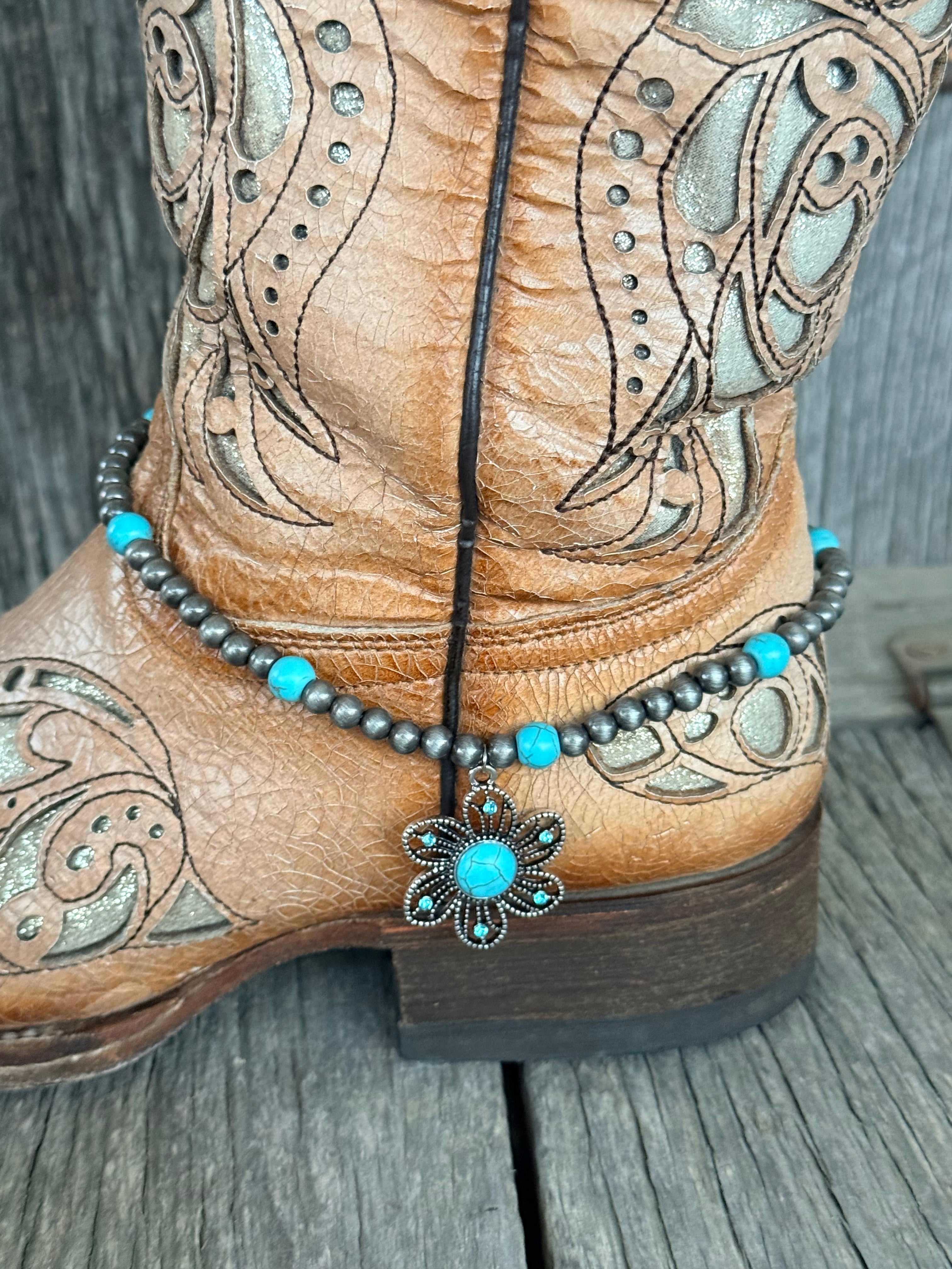 Side view of gunmetal Navajo pearl and turquoise bead boot bracelet with ornate silver flower concho pendant draping at ankle of tan tooled laser-cut cowboy boot on wood surface
