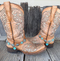 Turquoise flower concho chain boot bracelet with open silver heart and turquoise stone drop worn on tan tooled laser-cut cowboy boots, full pair front view on wood surface
