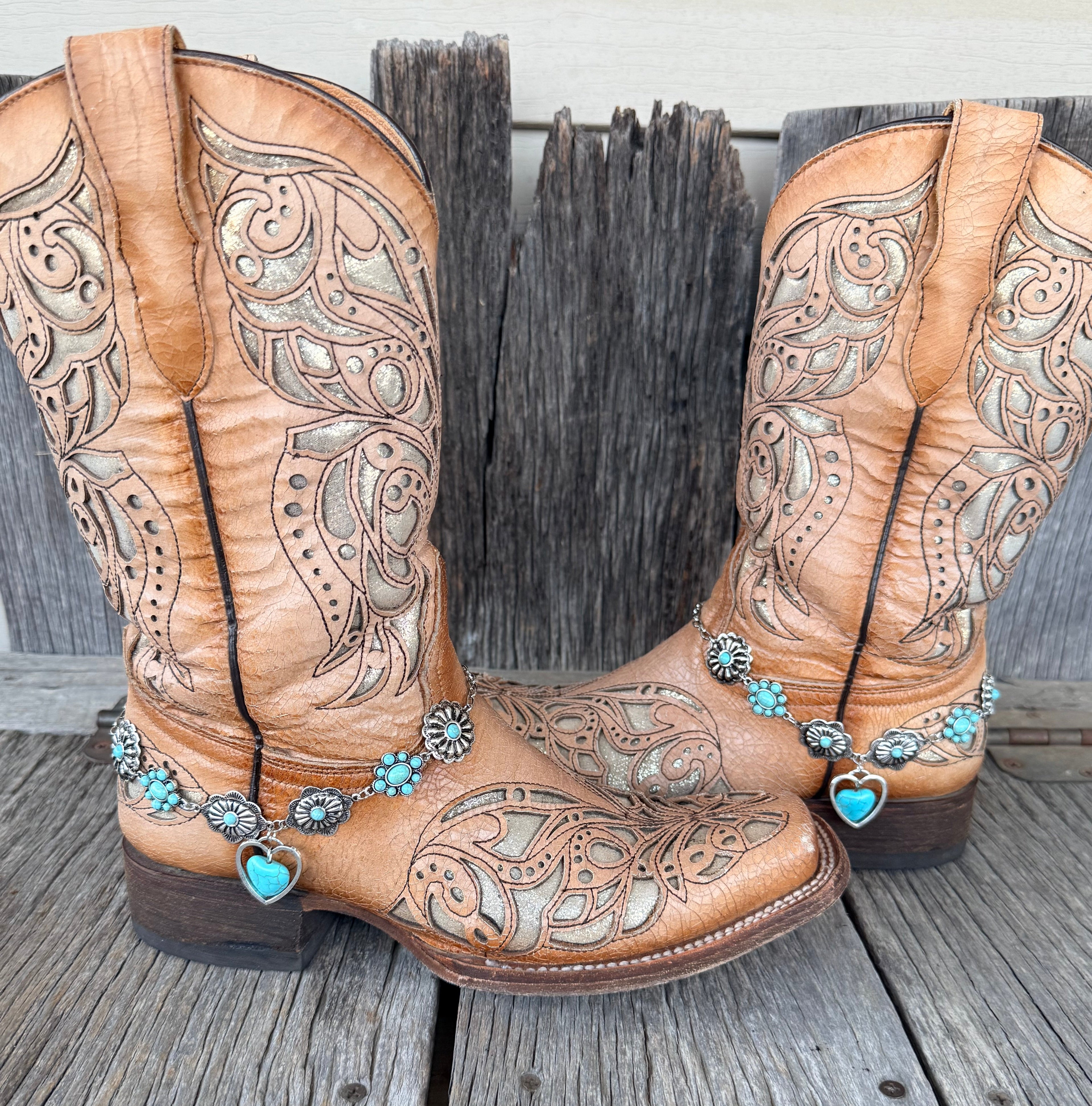 Turquoise flower concho chain boot bracelet with open silver heart and turquoise stone drop worn on tan tooled laser-cut cowboy boots, full pair front view on wood surface
