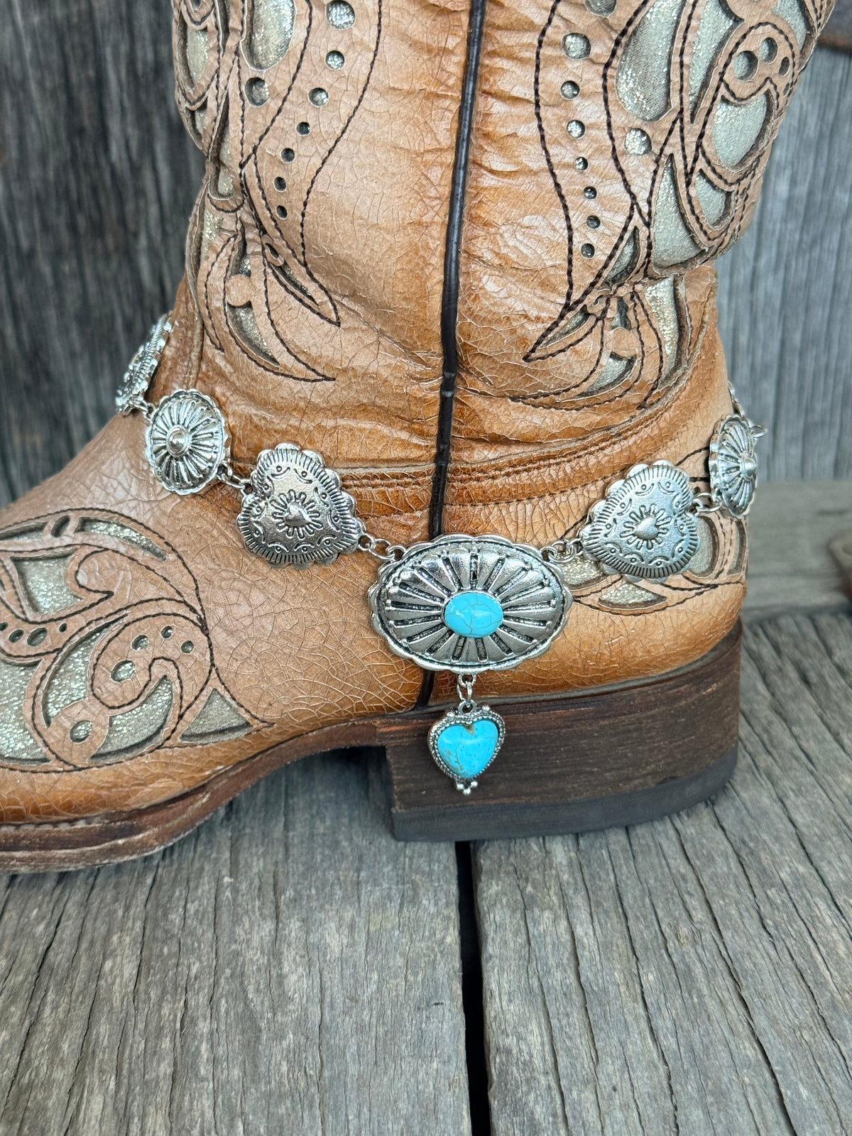 Side close-up of mixed stamped silver concho chain boot bracelet with oval turquoise centerpiece and heart pendant drop draping at ankle of tan laser-cut cowboy boot
