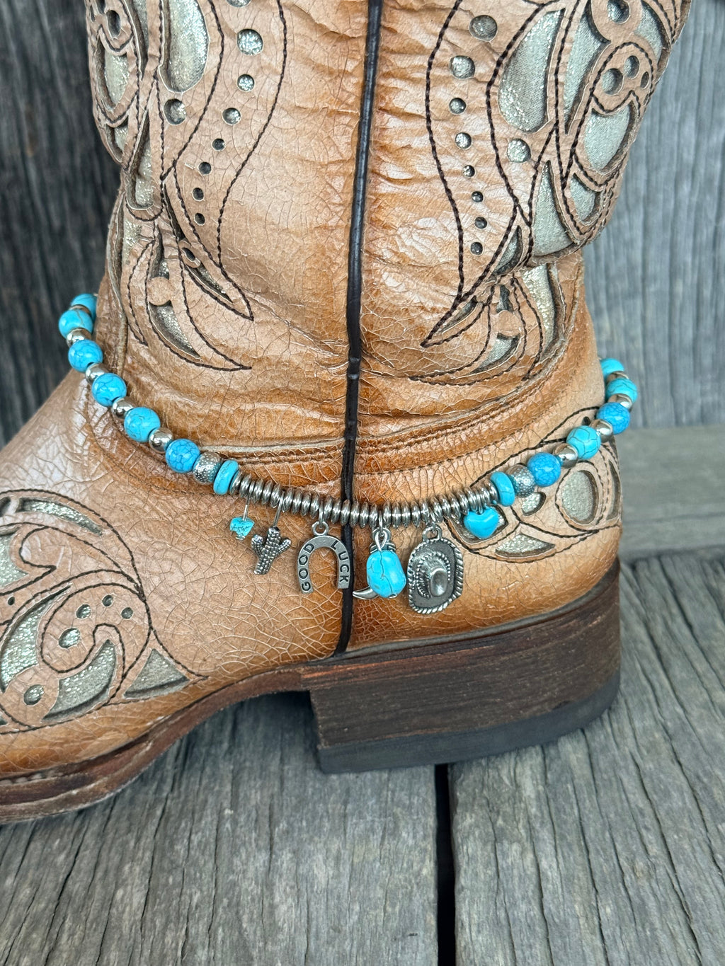 Side view of turquoise bead boot bracelet with Western silver charm cluster including Good Luck horseshoe, cactus, cowboy hat and cowboy boot charms draping at ankle of tan laser-cut tooled cowboy boot
