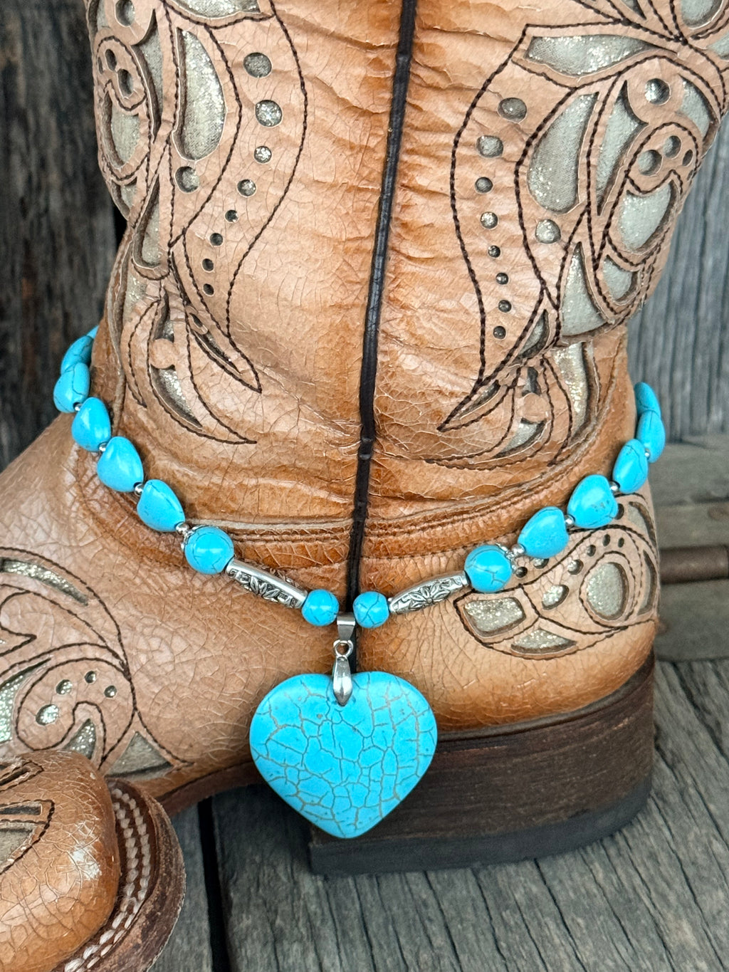 Back view of turquoise heart bead and round bead boot bracelet with large crackle turquoise heart stone pendant on silver bail at heel of tan tooled laser-cut cowboy boot on wood surface
