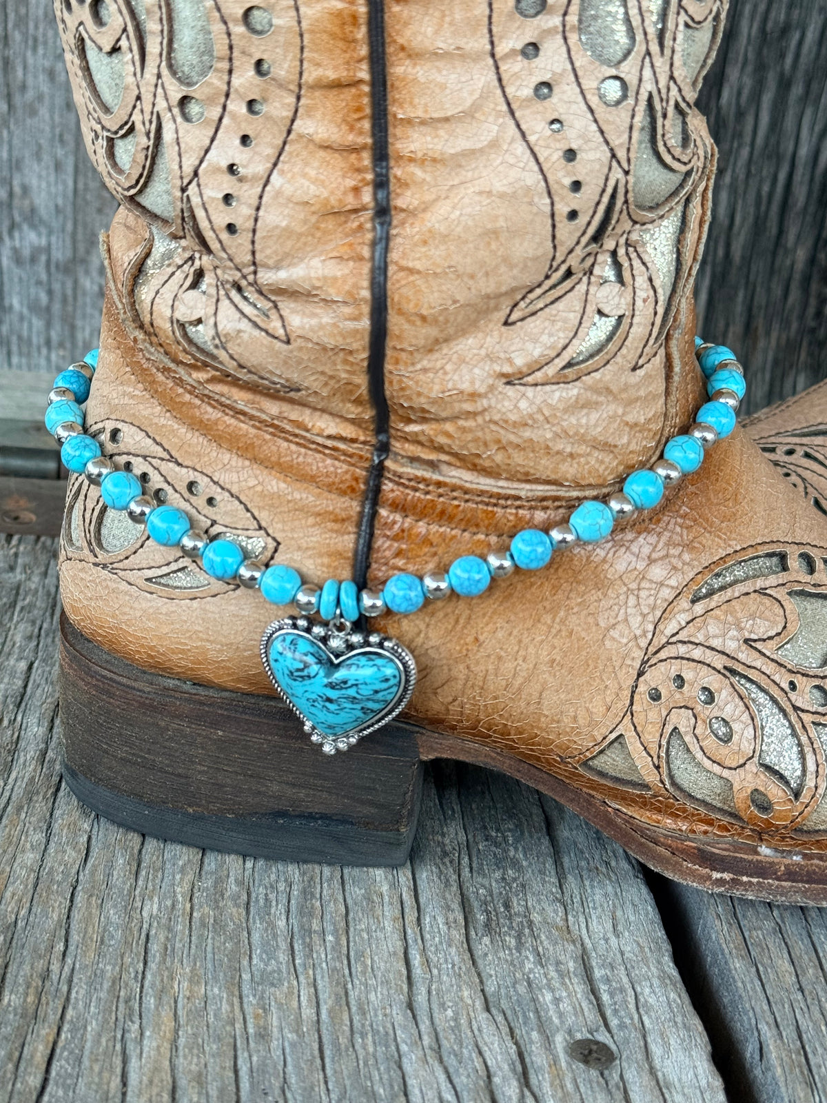 Close-up of turquoise heart pendant in silver dotted bezel on turquoise round bead boot bracelet at heel of tan cowboy boot

