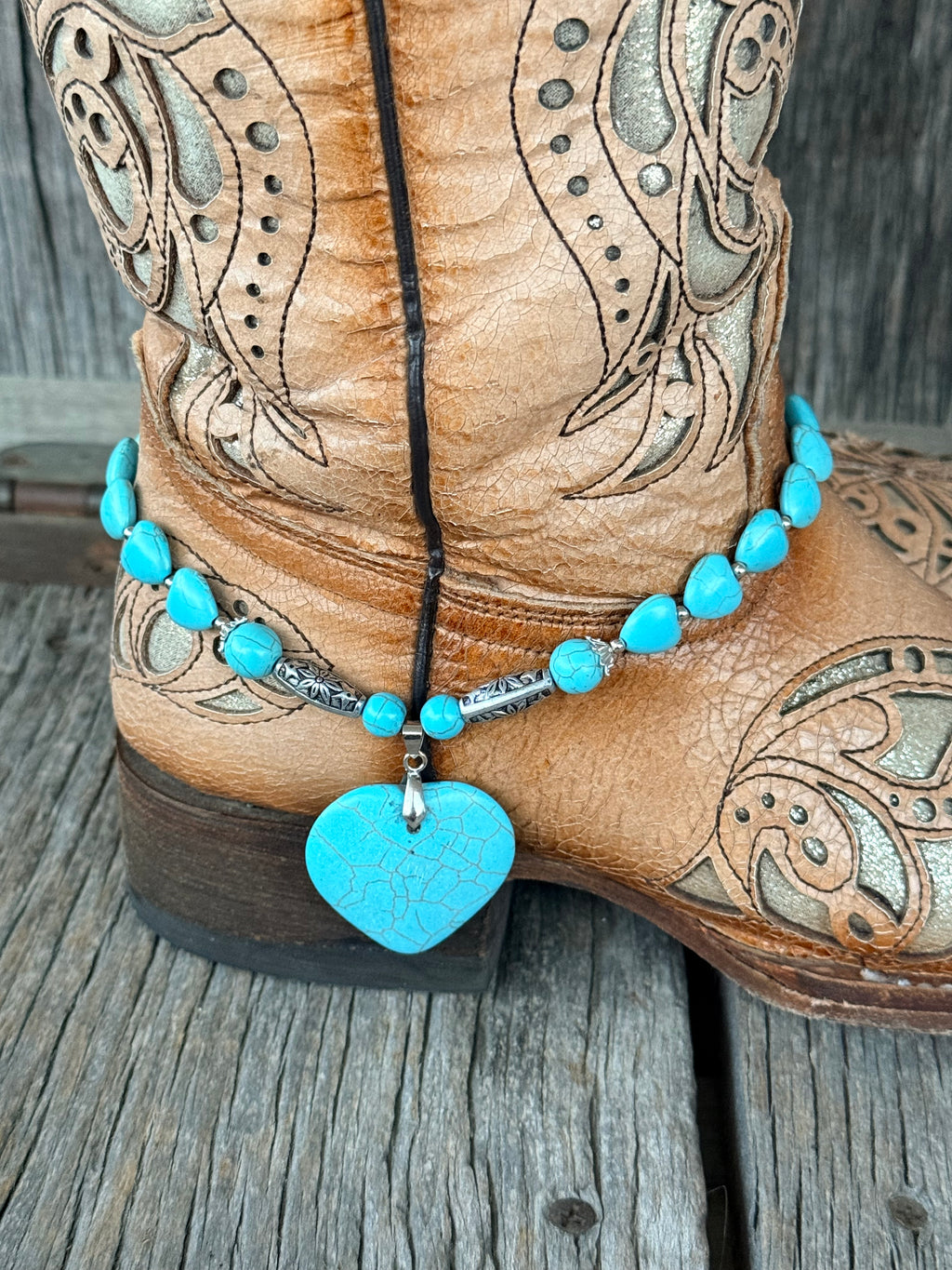 Close-up of turquoise heart bead and silver rondelle boot bracelet with engraved silver tube beads and large crackle turquoise heart stone pendant at heel of tan tooled cowboy boot
