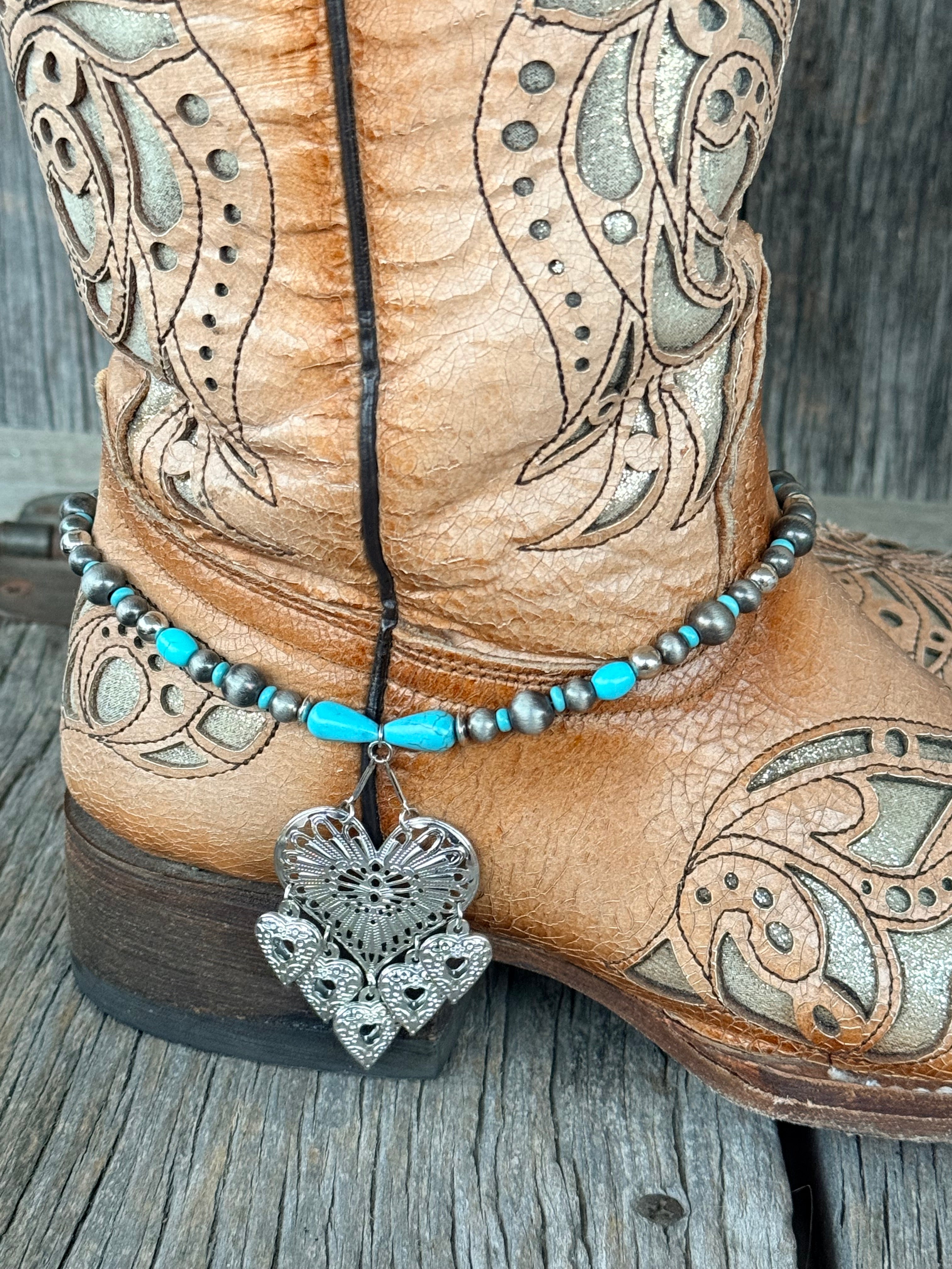 Close-up of gunmetal Navajo pearl and turquoise teardrop bead boot bracelet with large ornate silver filigree heart pendant and five dangling heart charms at heel of tan tooled cowboy boot
