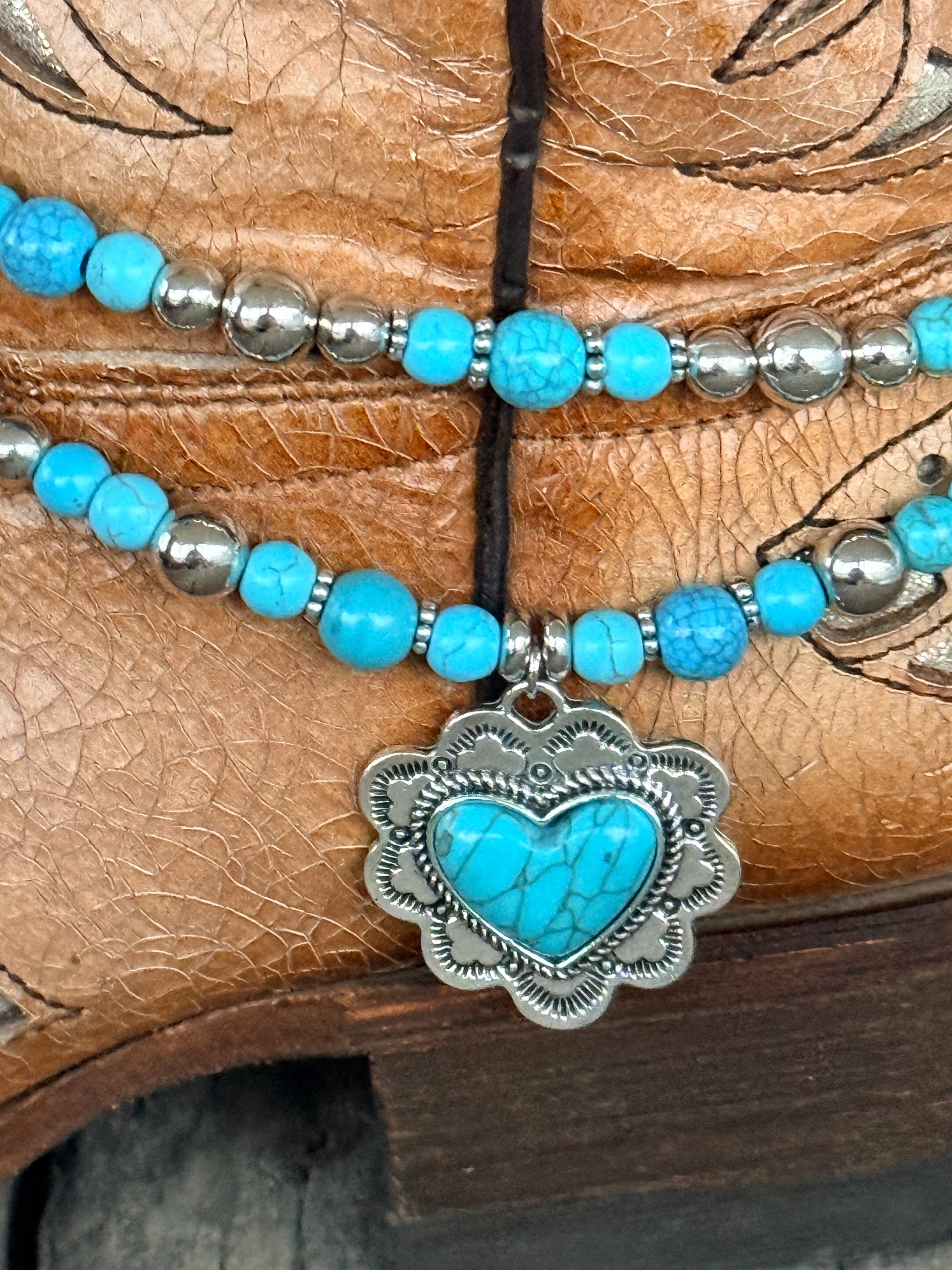 Detail close-up of ornate silver heart concho pendant with engraved scalloped border and turquoise heart cabochon center on double strand turquoise and silver round bead boot bracelet
