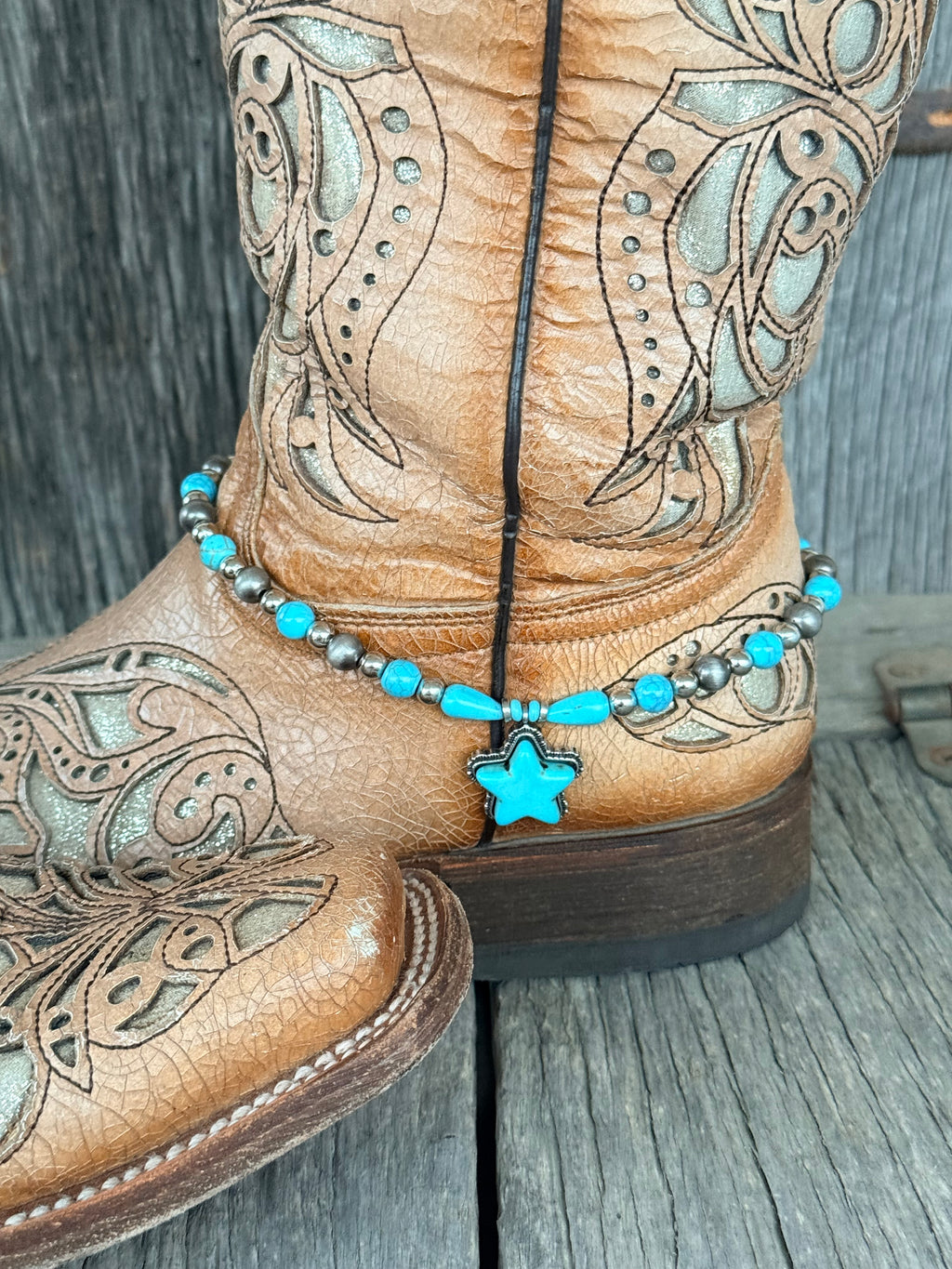 Side view of gunmetal Navajo pearl and turquoise bead boot bracelet with turquoise teardrop beads and large turquoise star stone pendant draping at ankle of tan tooled laser-cut cowboy boot on wood surface
