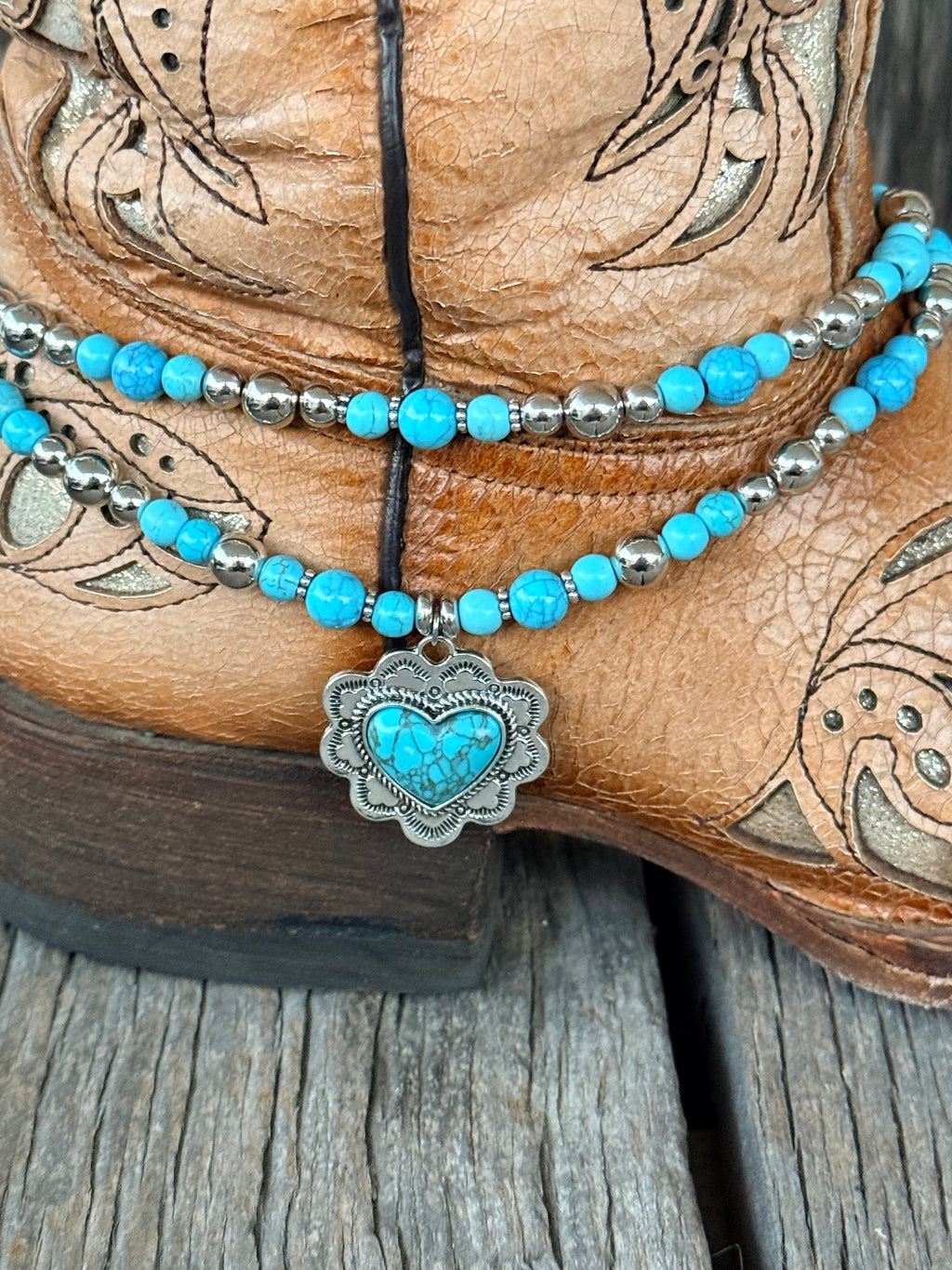 Side close-up of double strand turquoise and silver bead boot bracelet with ornate engraved silver heart concho pendant and turquoise heart cabochon center draping at ankle of tan tooled cowboy boot

