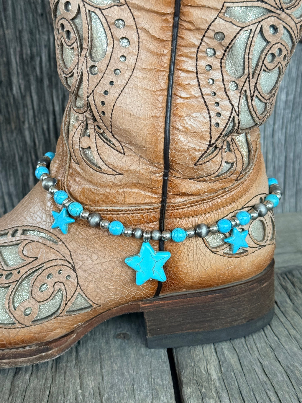 Side view of Navajo pearl and turquoise bead boot bracelet with three turquoise star stone pendants draping at ankle of tan tooled laser-cut cowboy boot on wood surface
