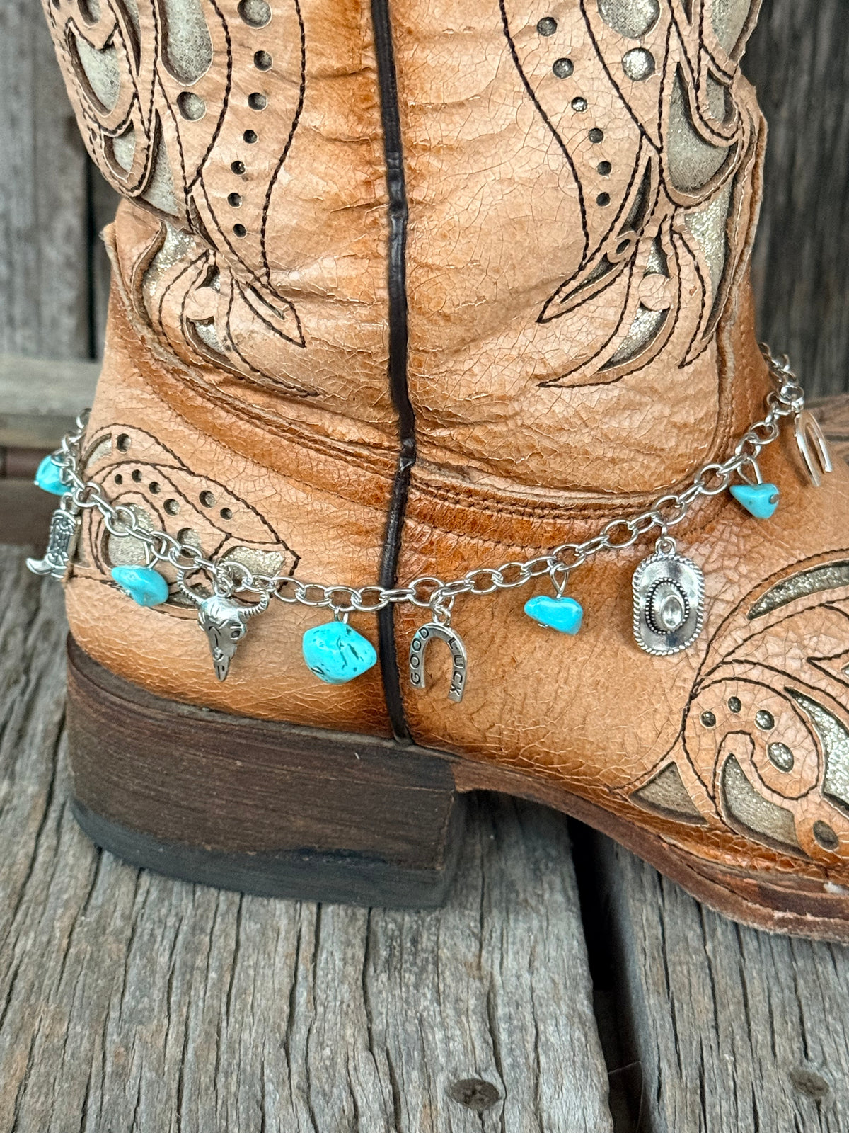 Close-up of silver curb chain boot bracelet with turquoise stone chips and silver steer head and Good Luck horseshoe charms at heel of tan tooled cowboy boot on wood surface
