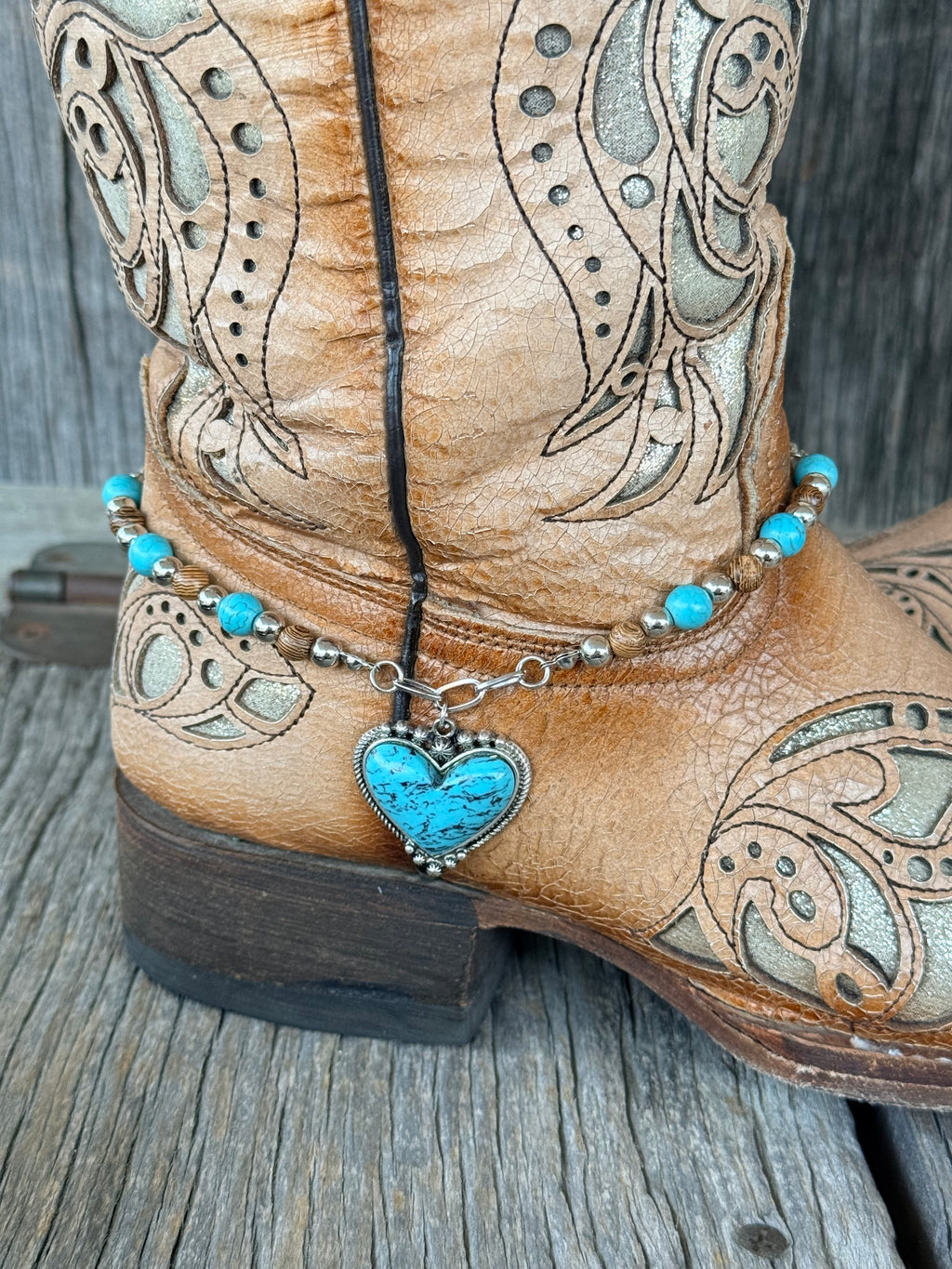 Close-up of turquoise heart pendant in silver filigree bezel dropping from toggle ring on beaded boot bracelet at heel of tan cowboy boot

