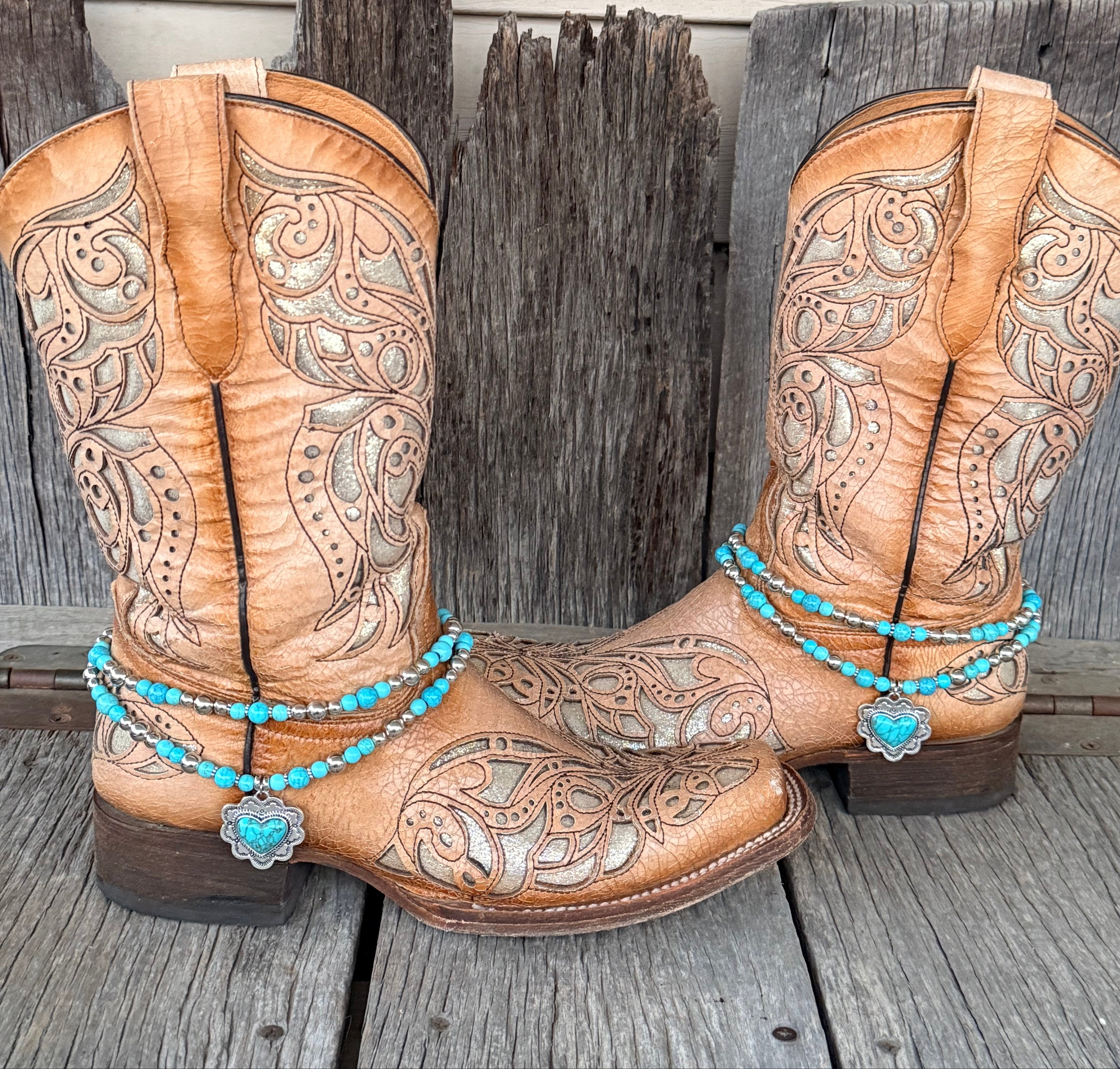 Double strand turquoise and silver bead boot bracelet with ornate silver heart concho pendant worn on tan tooled laser-cut cowboy boots, full pair front view against weathered wood
