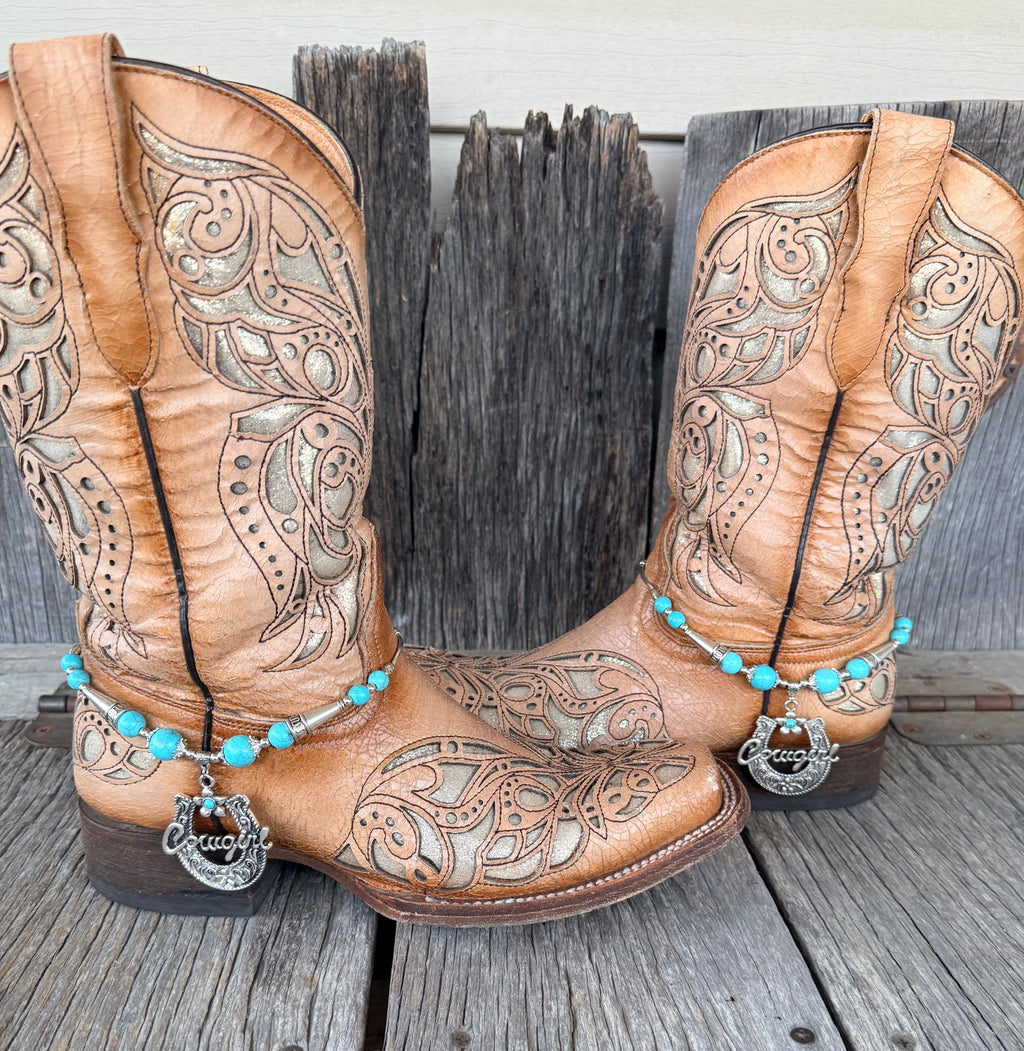 Turquoise and silver Cowgirl horseshoe boot bracelet with script pendant worn on tan tooled laser-cut cowboy boots, full pair front view on wood surface
