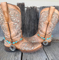 Turquoise and silver Cowgirl horseshoe boot bracelet with script pendant worn on tan tooled laser-cut cowboy boots, full pair front view on wood surface
