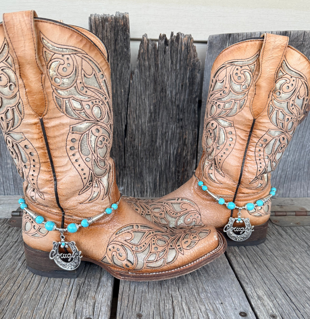 Turquoise and silver Cowgirl horseshoe boot bracelet with script pendant worn on tan tooled laser-cut cowboy boots, full pair front view on wood surface
