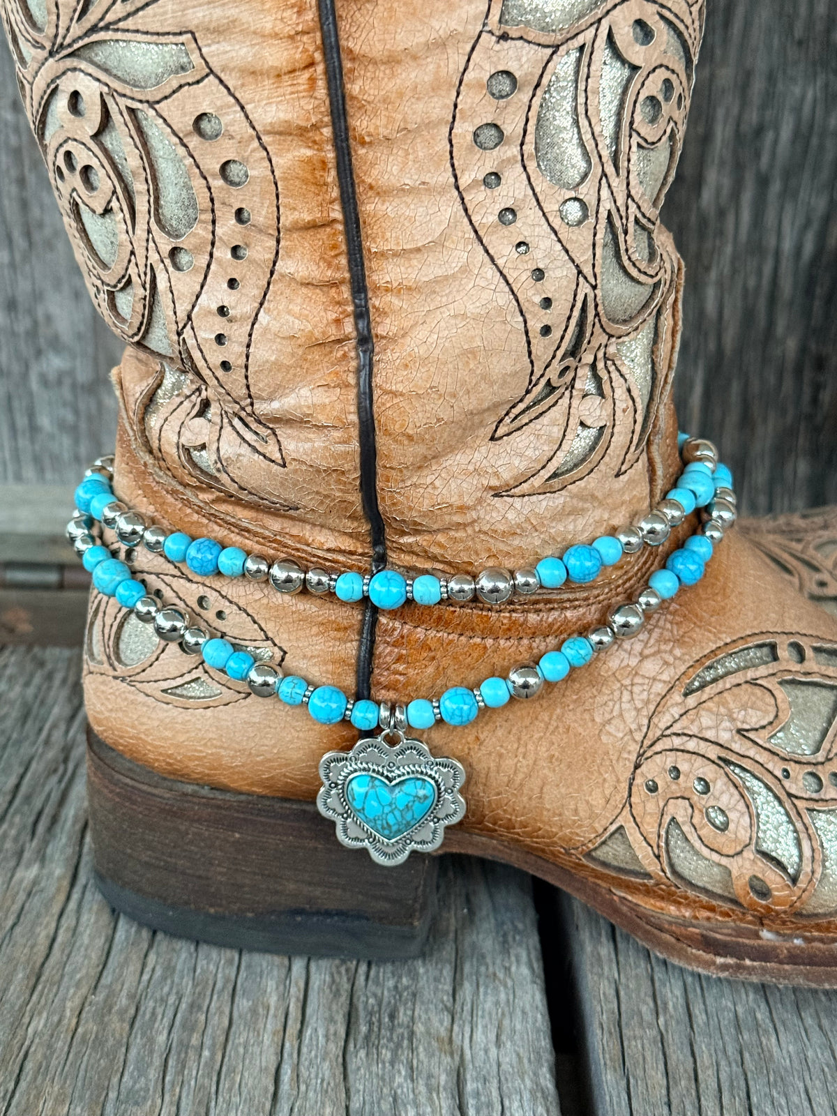 Close-up of double strand turquoise round bead and silver round bead boot bracelet with ornate silver heart concho pendant and turquoise heart cabochon at heel of tan tooled cowboy boot
