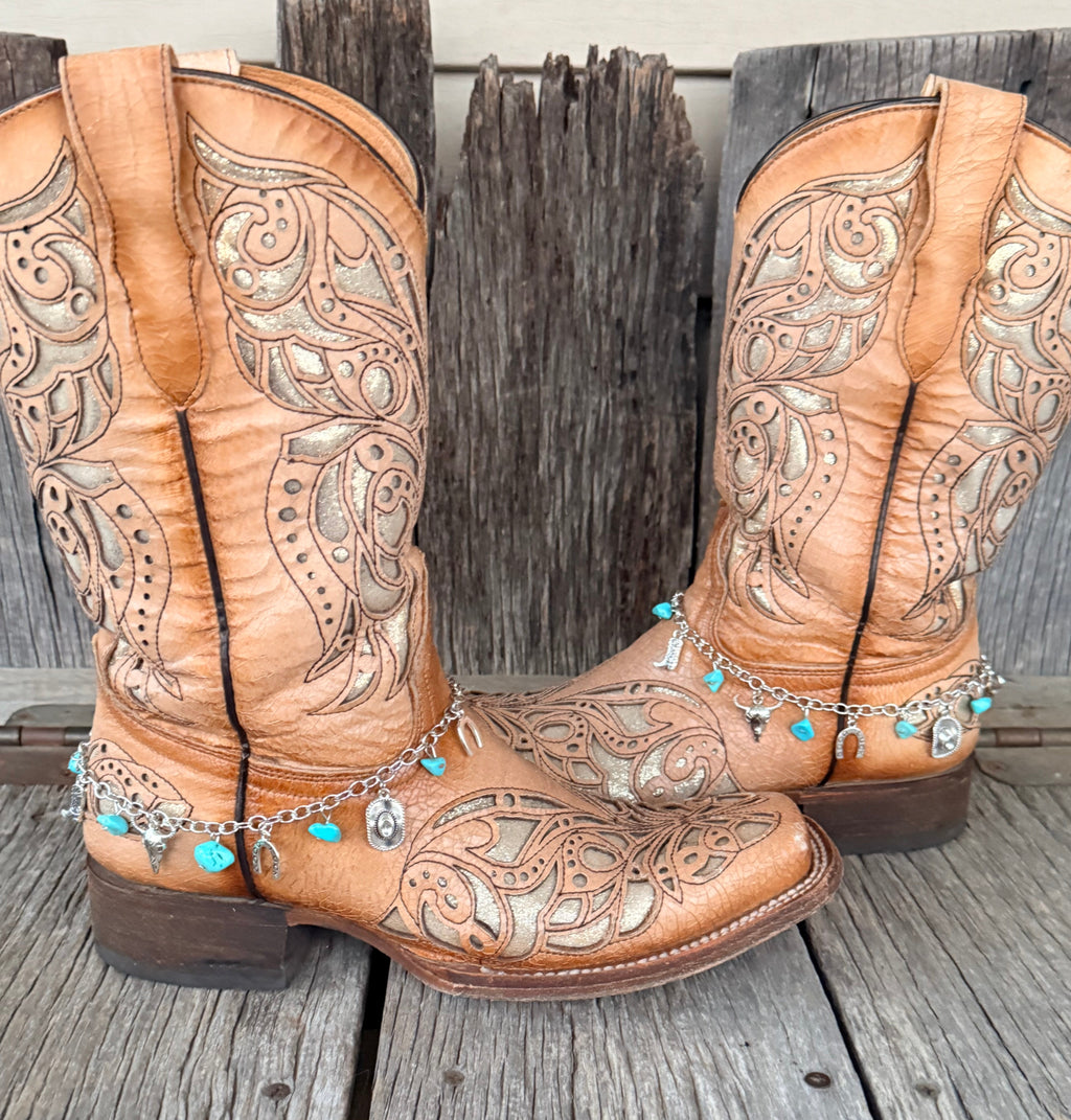 Western charm boot bracelet with turquoise stone chips and silver cowboy boot, steer head, Good Luck horseshoe, and cowboy hat charms worn on tan tooled laser-cut cowboy boots, full pair front view against weathered wood
