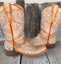Western charm boot bracelet with turquoise stone chips and silver cowboy boot, steer head, Good Luck horseshoe, and cowboy hat charms worn on tan tooled laser-cut cowboy boots, full pair front view against weathered wood
