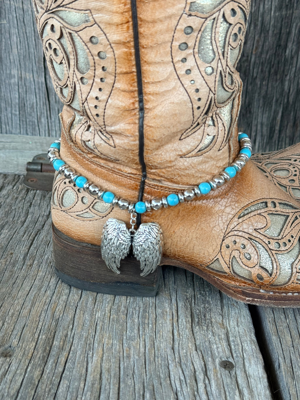 Close-up of large silver angel wing pair pendant dropping from turquoise and silver beaded boot bracelet at heel of tan tooled leather cowboy boot
