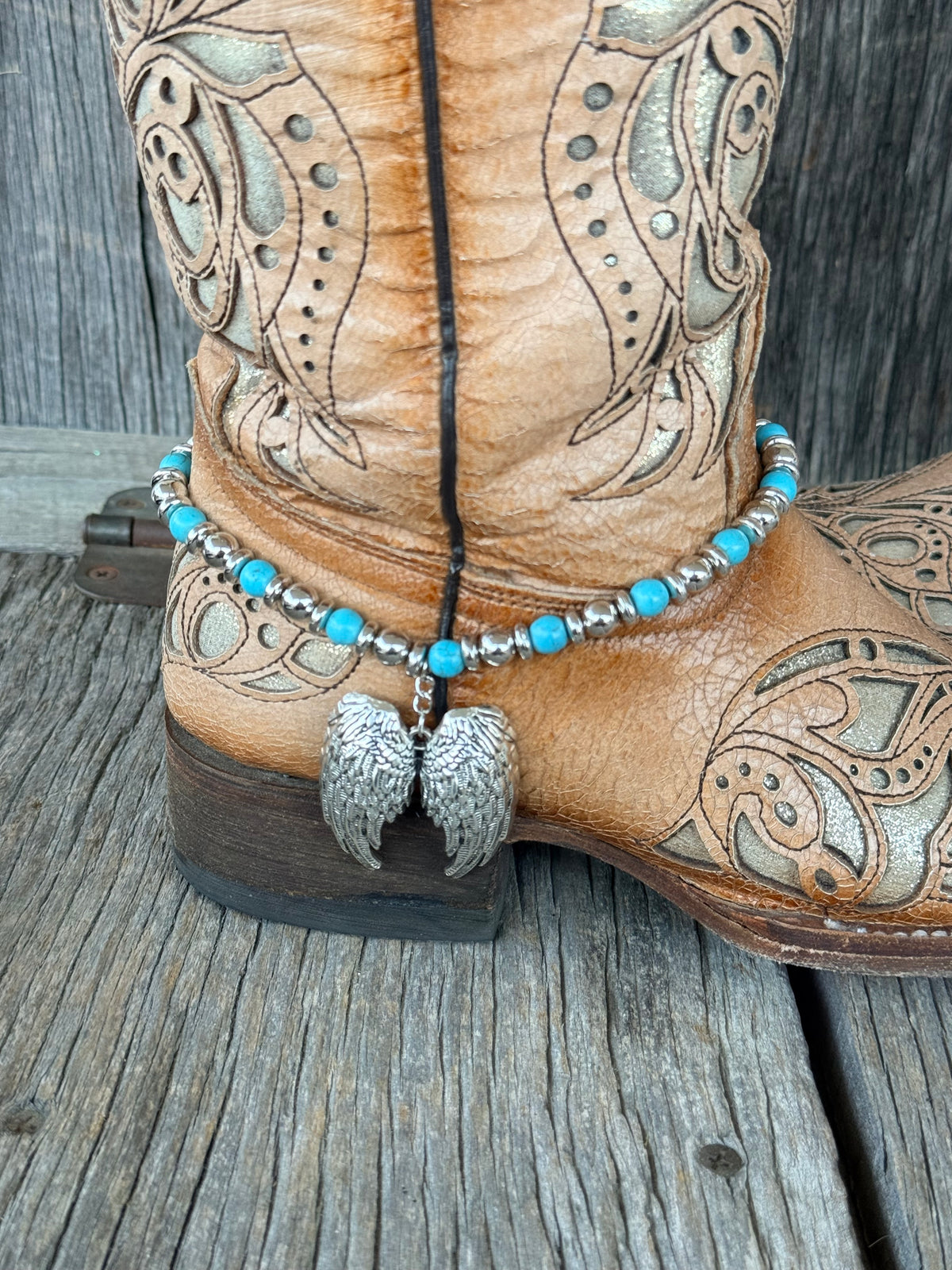 Close-up of large silver angel wing pair pendant dropping from turquoise and silver beaded boot bracelet at heel of tan tooled leather cowboy boot
