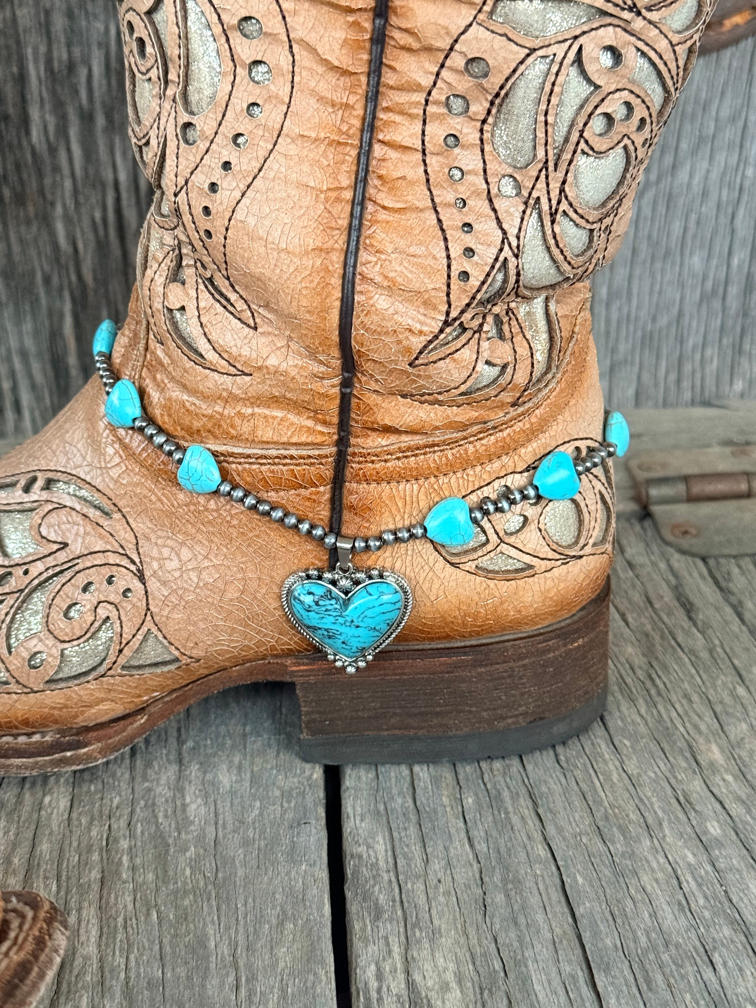 Side view of turquoise heart bead and Navajo pearl gunmetal bead boot bracelet with large turquoise heart stone pendant hanging at ankle of tan tooled laser-cut cowboy boot on wood surface
