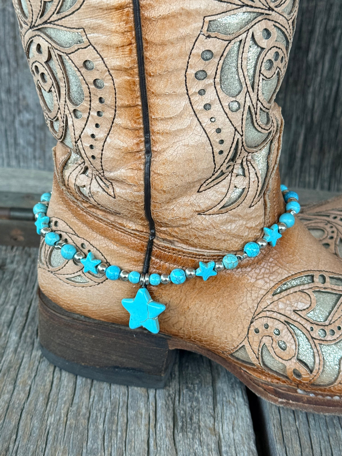 Close-up of turquoise round bead and silver rondelle spacer boot bracelet with multiple turquoise star beads along strand and large turquoise star stone pendant drop at heel of tan tooled cowboy boot

