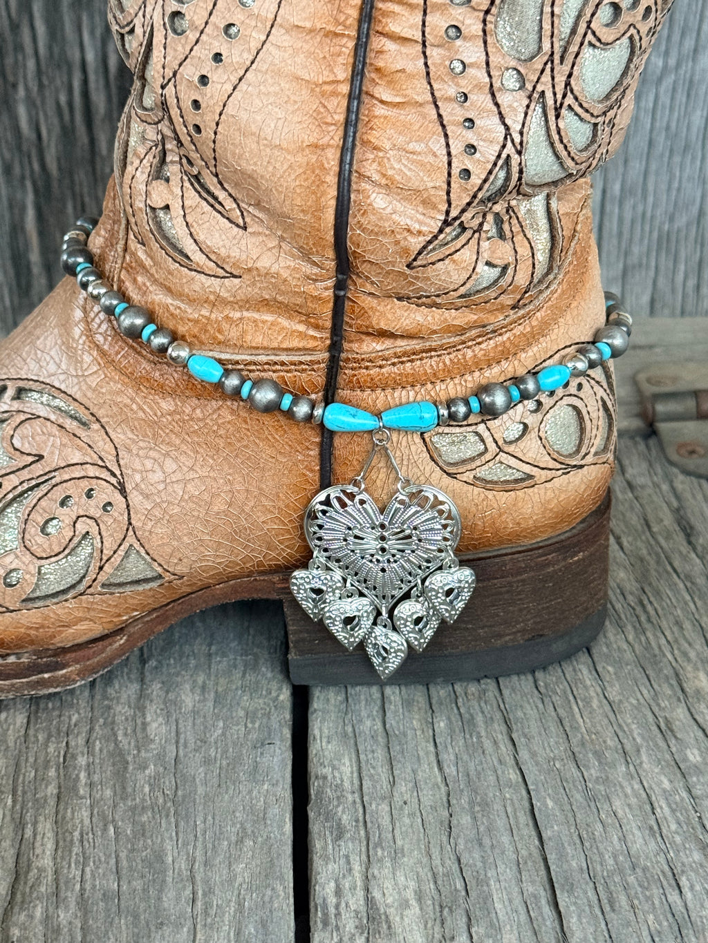 Low angle front view of turquoise teardrop and Navajo pearl boot bracelet with large ornate silver filigree heart pendant and five dangling heart charms at heel of tan tooled cowboy boot on wood surface
