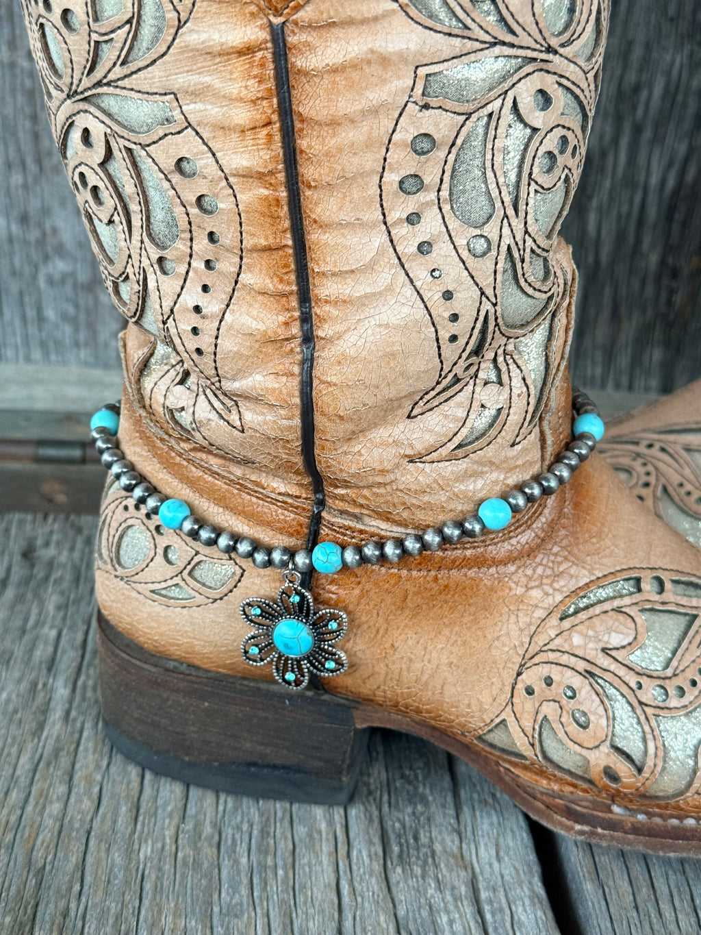 Close-up of gunmetal Navajo pearl and turquoise bead boot bracelet with large ornate silver flower concho pendant with turquoise cabochon center at heel of tan tooled cowboy boot
