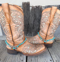 Turquoise bead and silver Good Luck charm boot bracelet with Western charm cluster worn on tan tooled laser-cut cowboy boots, full pair front view on wood surface
