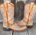Turquoise heart bead and heart stone boot bracelet with large turquoise heart pendant worn on tan tooled laser-cut cowboy boots, full pair front view against weathered wood fence
