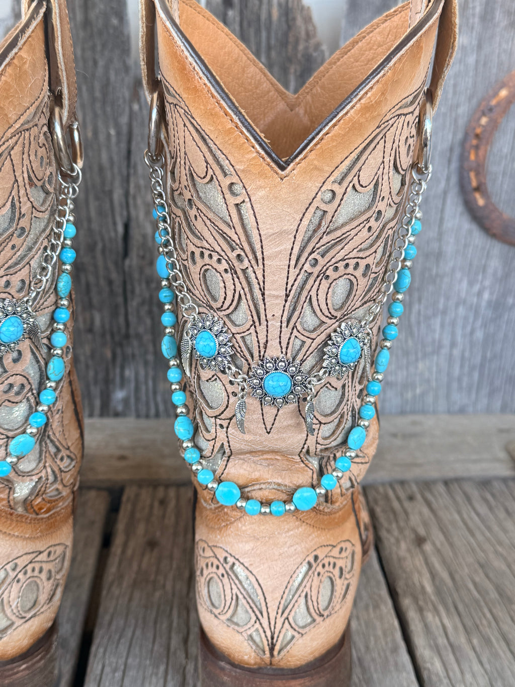 Side angle of silver flower concho and turquoise bead boot bracelet chain draped across shaft of tan tooled cowboy boots, full pair view
