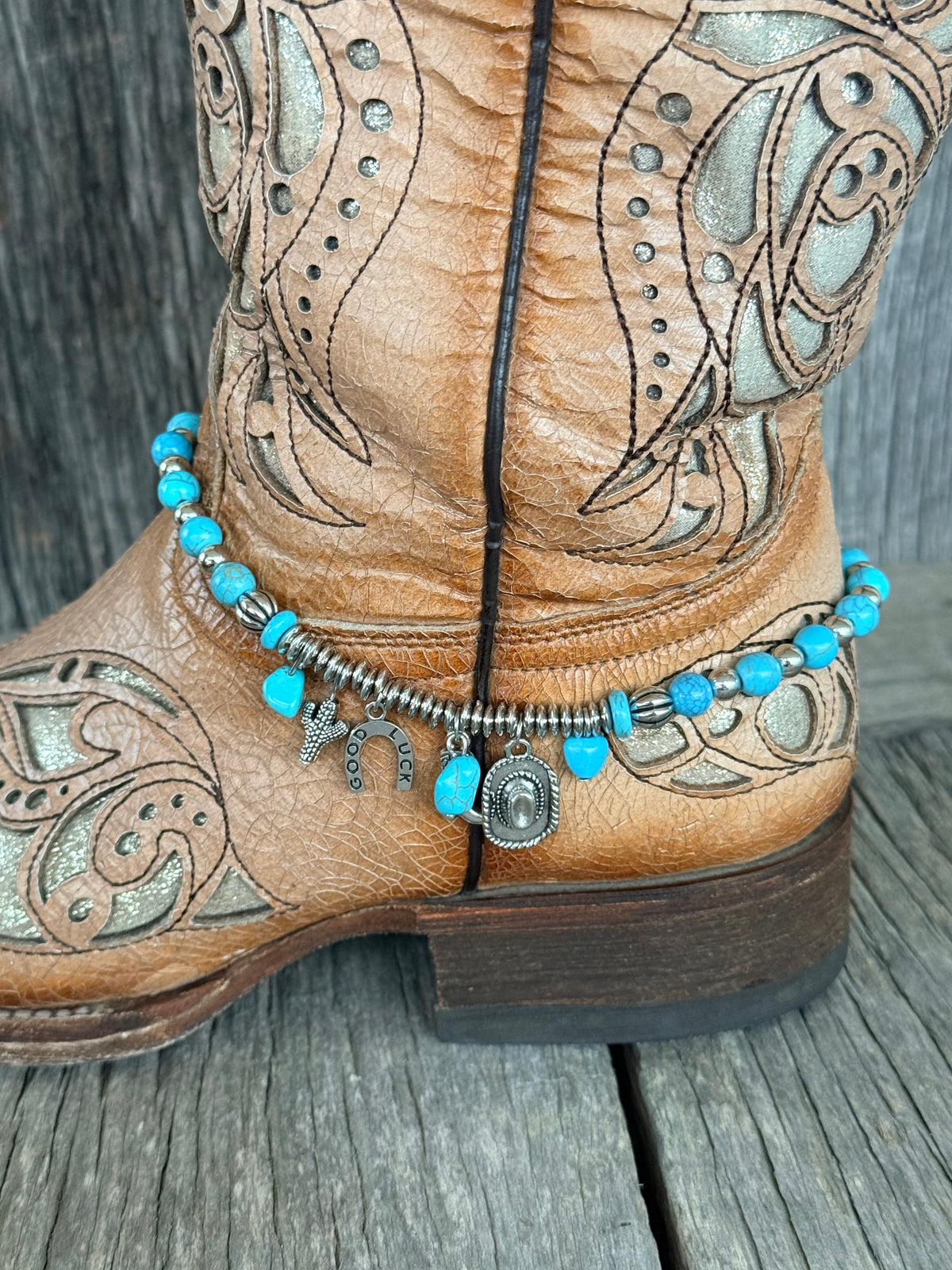 Back view close-up of turquoise bead boot bracelet with silver ribbed barrel spacers and Western charm cluster with Good Luck horseshoe and cowboy hat pendant at heel of tan tooled cowboy boot
