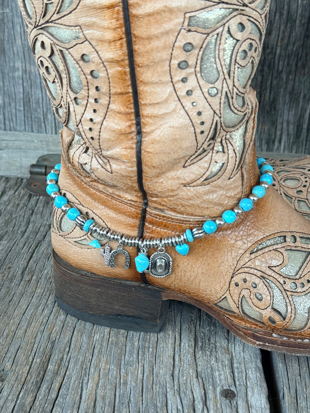 Side close-up of turquoise bead and silver ribbed barrel spacer boot bracelet with Good Luck horseshoe, cactus, and cowboy hat charm cluster draping at ankle of tan tooled laser-cut cowboy boot on wood surface
