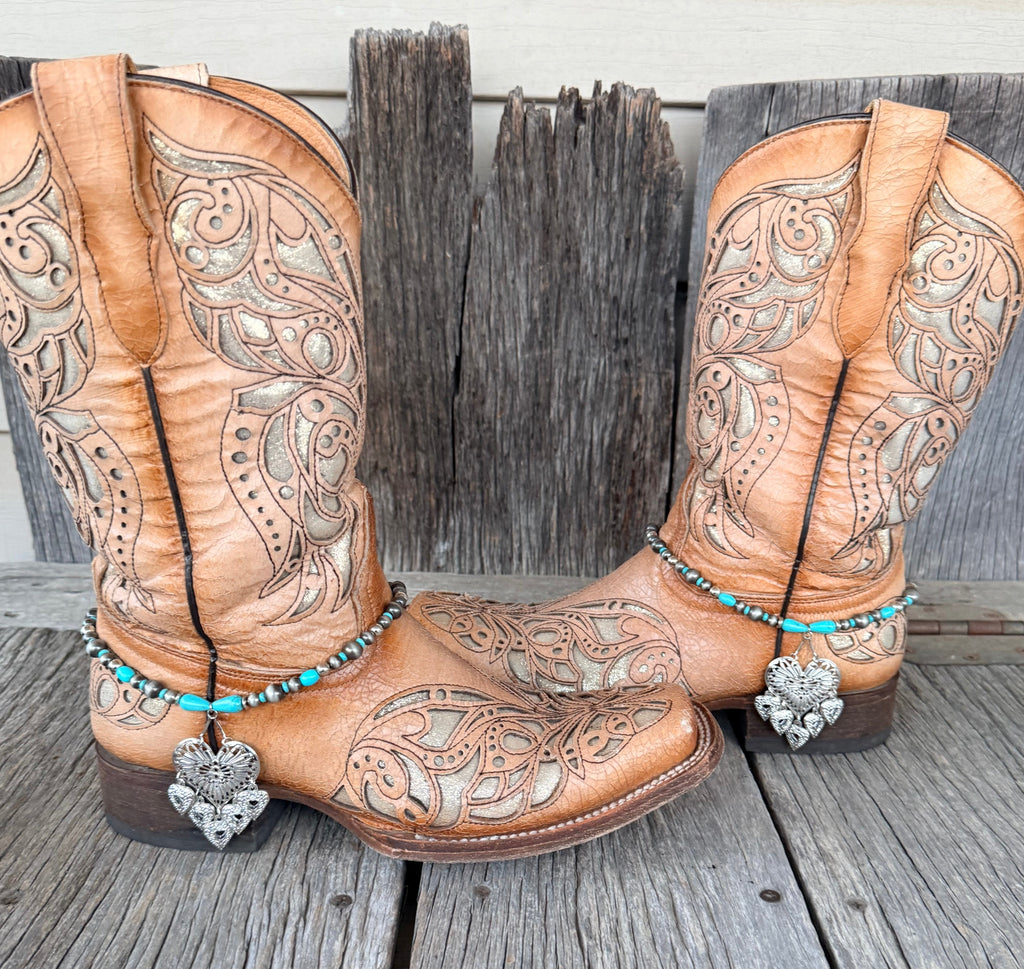 Silver filigree heart and turquoise teardrop bead boot bracelet with large ornate heart pendant worn on tan tooled laser-cut cowboy boots, full pair front view on wood surface

