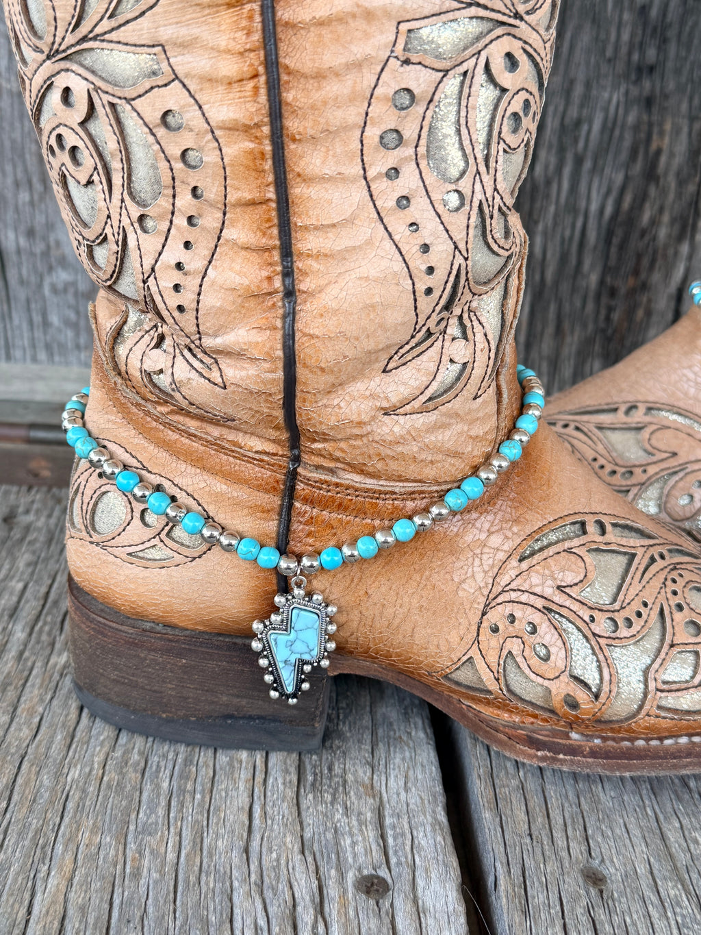 Close-up of turquoise lightning bolt pendant in silver bezel with ball bead trim on beaded boot bracelet at heel of tan cowboy boot
