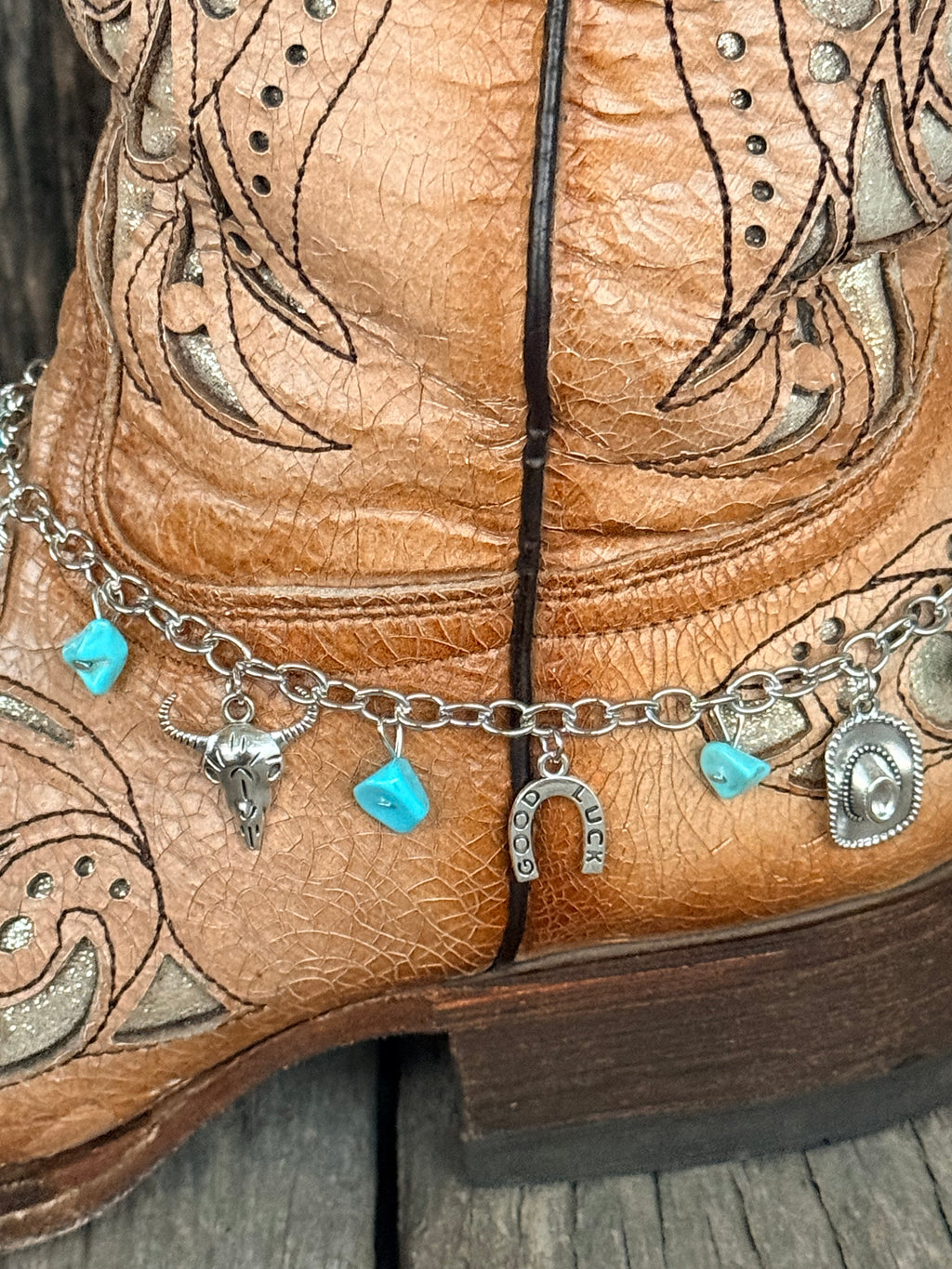 Front detail close-up of silver Western charm boot bracelet showing turquoise stone chips, silver steer head charm, Good Luck horseshoe charm, and cowboy hat charm on silver curb chain at ankle of tan tooled cowboy boot
