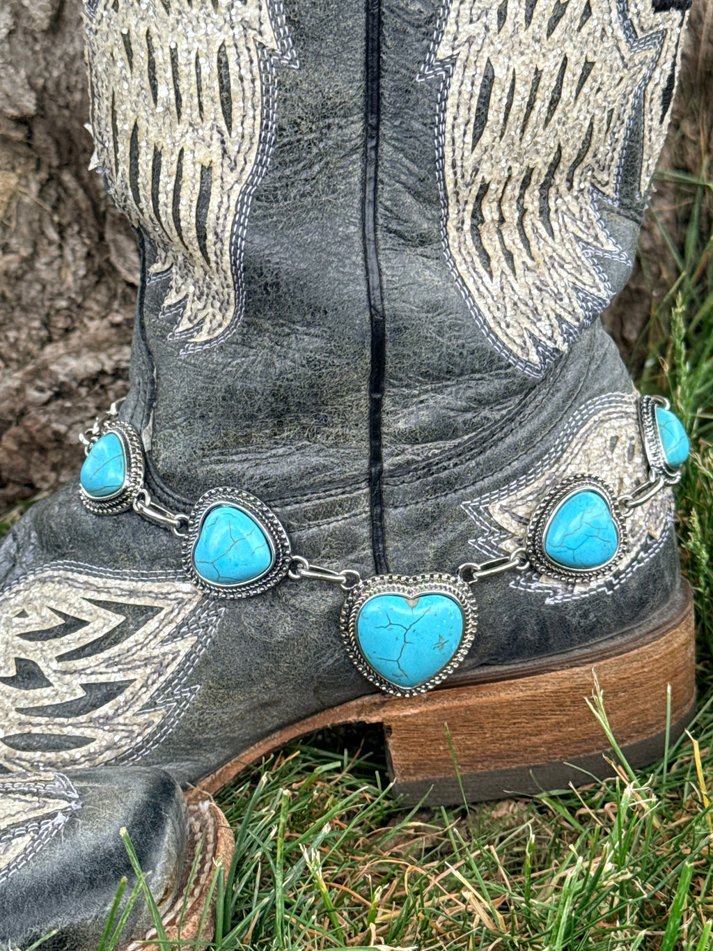 Side view of turquoise heart boot bracelet with five graduating heart-shaped howlite stones in silver beaded bezels draped across the toe box of a black angel wing embroidered cowboy boot on grass
