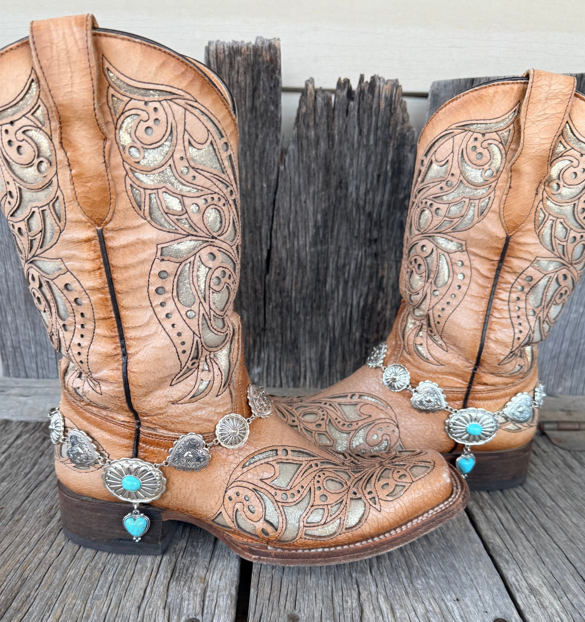 Silver concho chain boot bracelet with turquoise heart pendant worn on tan tooled laser-cut cowboy boots, full pair front view on wood surface
