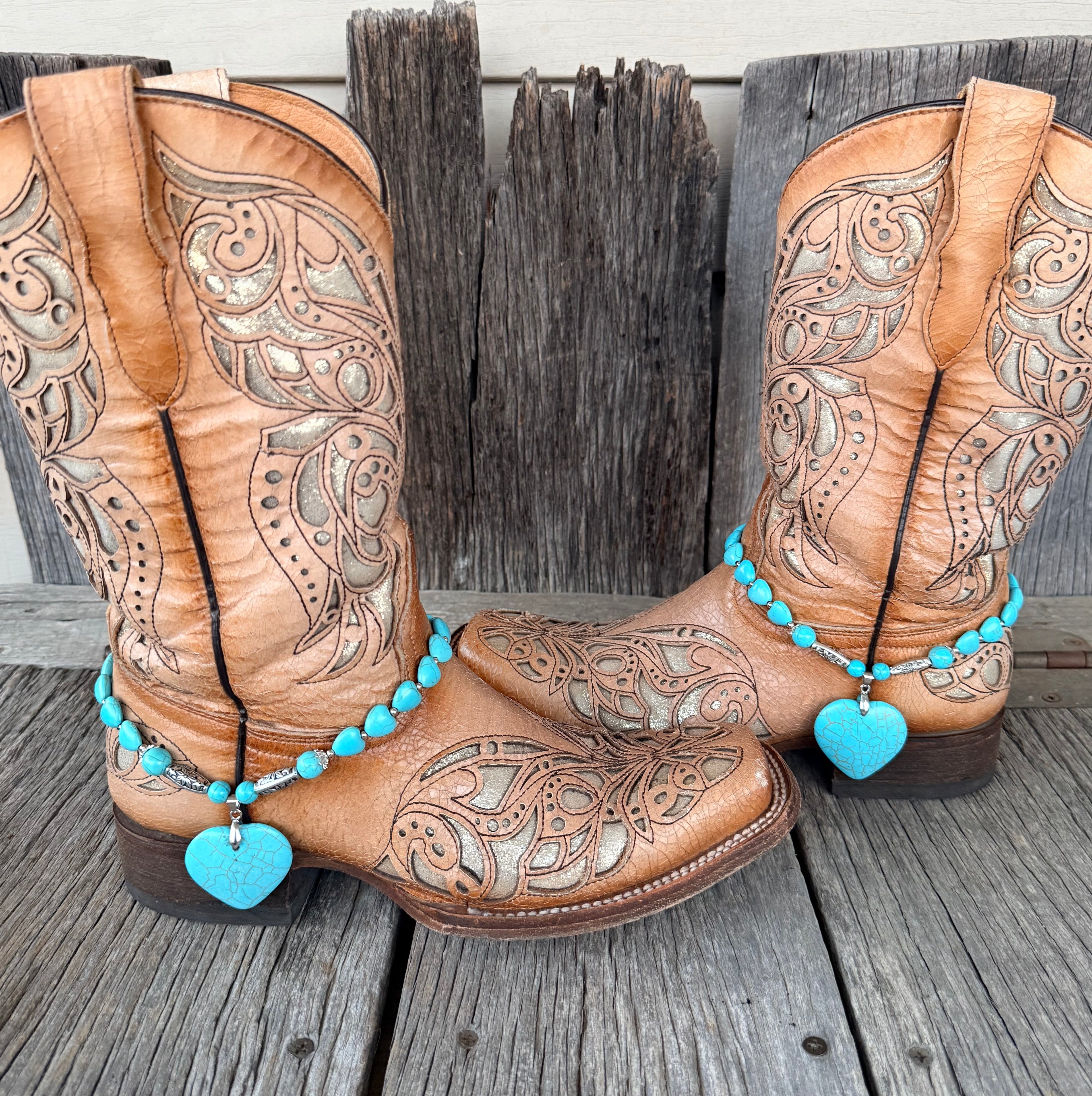Turquoise heart bead boot bracelet with large crackle turquoise heart stone pendant worn on tan tooled laser-cut cowboy boots, full pair front view against weathered wood
