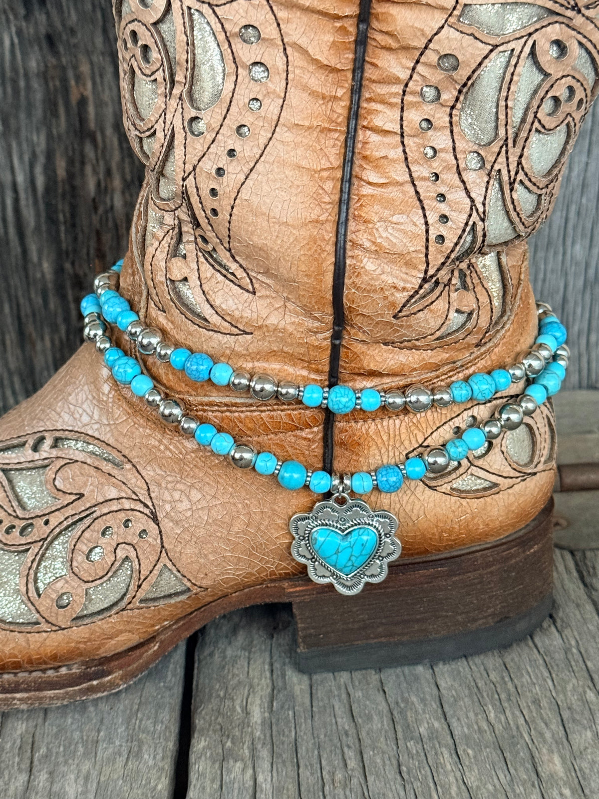 Side view of double strand turquoise round bead and silver bead boot bracelet with ornate silver heart concho pendant draping at ankle of tan tooled laser-cut cowboy boot on wood surface
