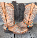 Turquoise and gunmetal Navajo pearl boot bracelet with ornate silver flower concho pendant worn on tan tooled laser-cut cowboy boots, full pair front view on wood surface

