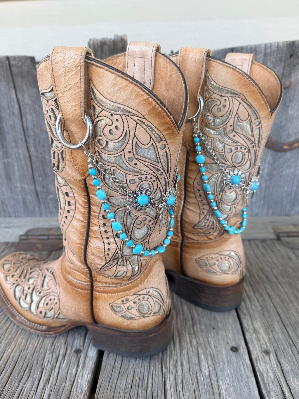 Side view of two-strand turquoise bead and silver chain boot bracelet with flower concho centerpiece draped across tan tooled cowboy boots on wood surface
