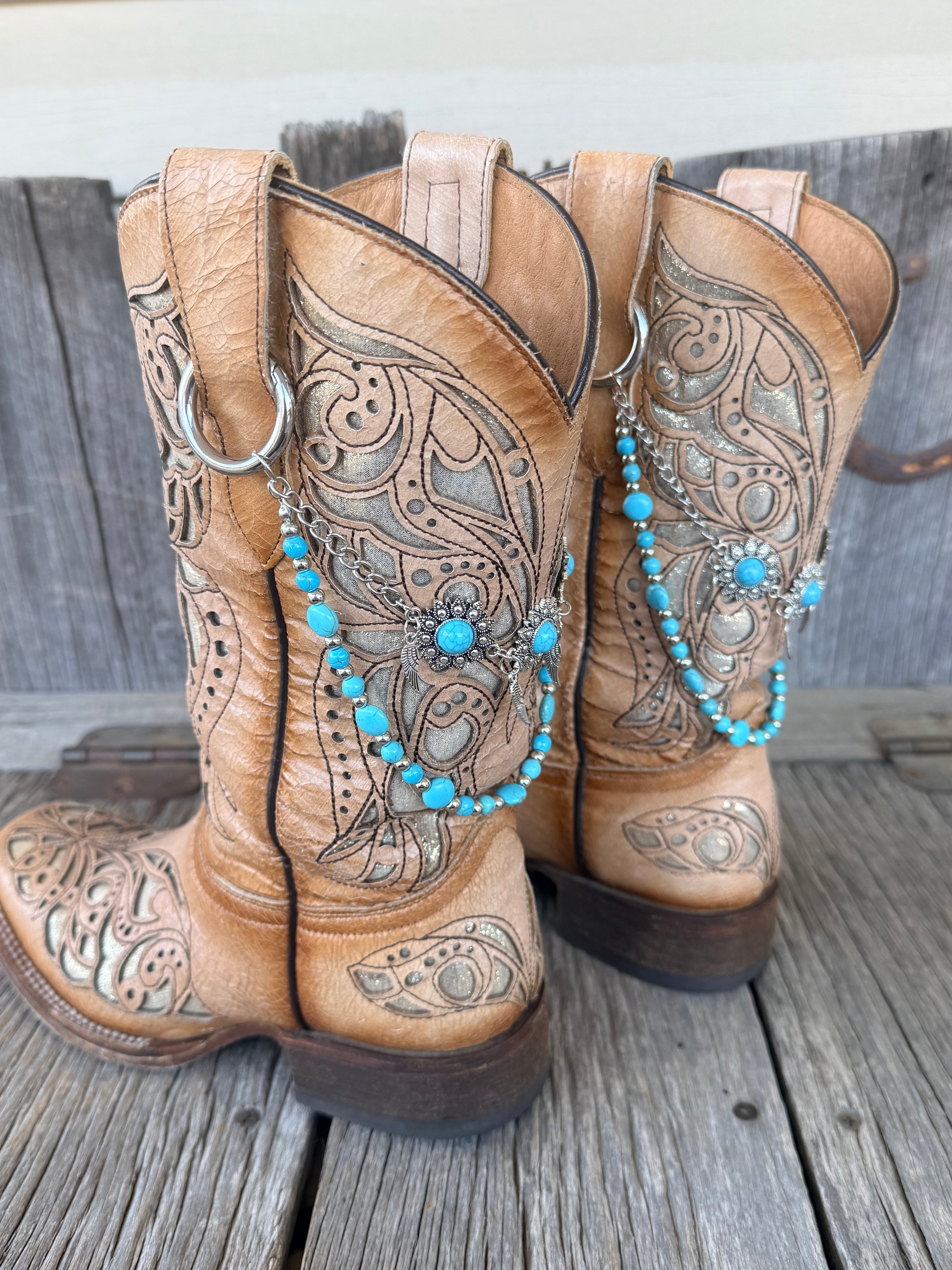 Side view of two-strand turquoise bead and silver chain boot bracelet with flower concho centerpiece draped across tan tooled cowboy boots on wood surface
