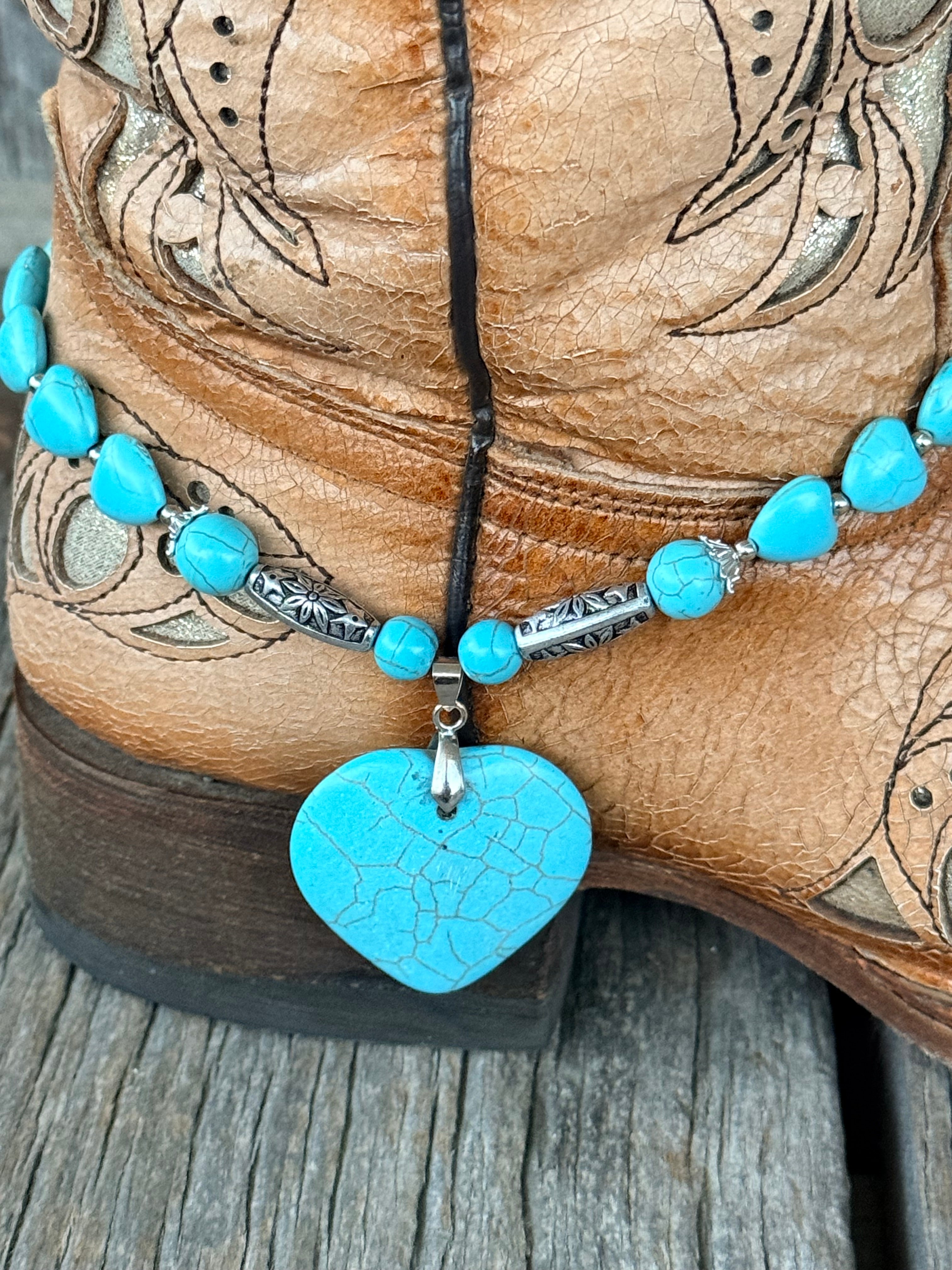 Side close-up of turquoise heart bead boot bracelet with engraved silver floral-stamped tube beads and large crackle turquoise heart stone pendant draping at ankle of tan tooled cowboy boot
