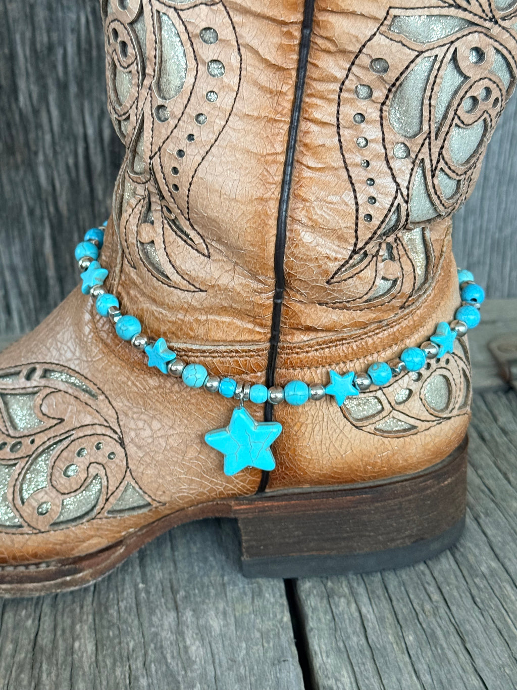 Side view of turquoise star beaded boot bracelet with silver rondelle spacers and large center turquoise star pendant draping at ankle of tan tooled laser-cut cowboy boot on wood surface
